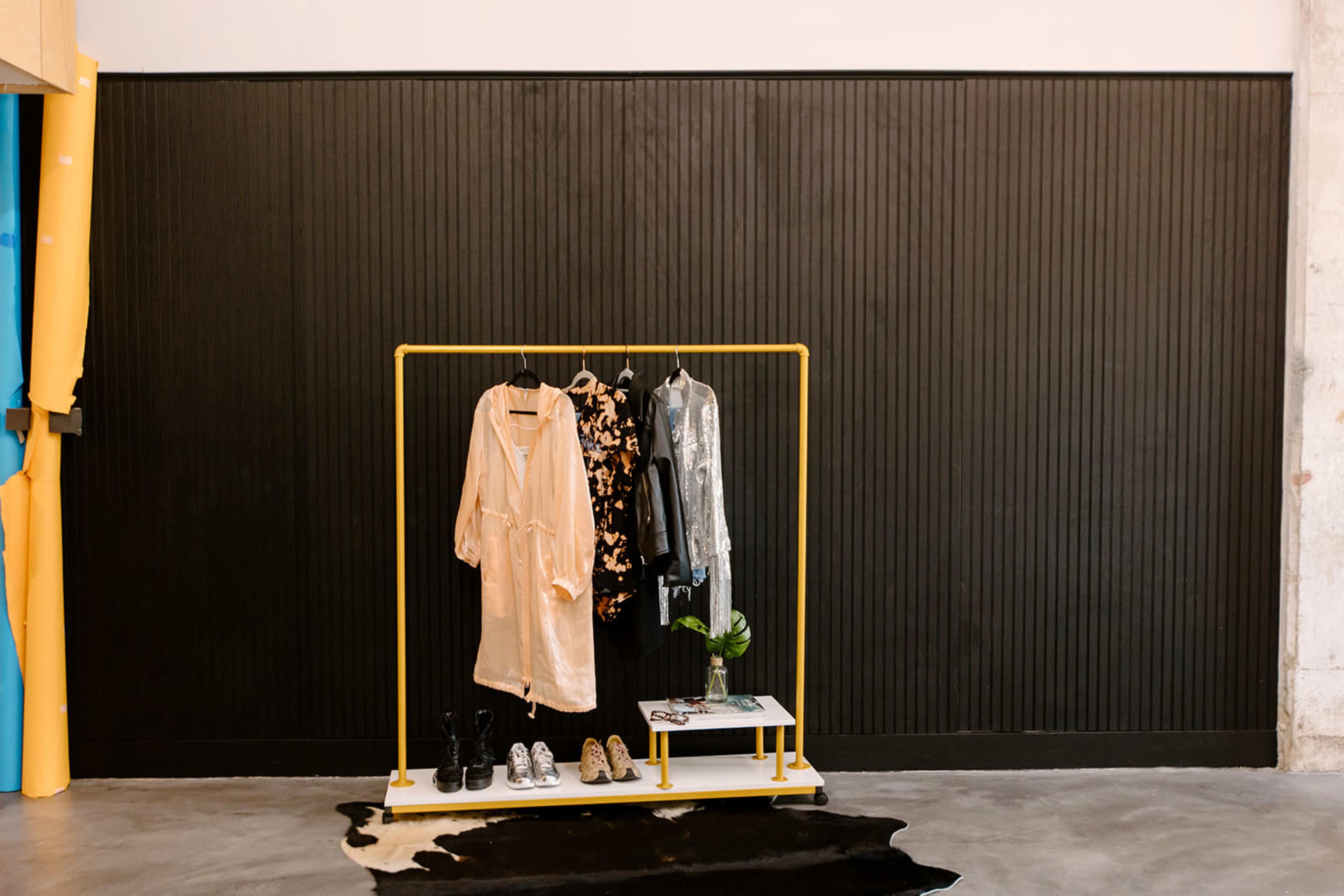 A clothing rack displaying two outfits and a pair of shoes is positioned against a black textured wall.