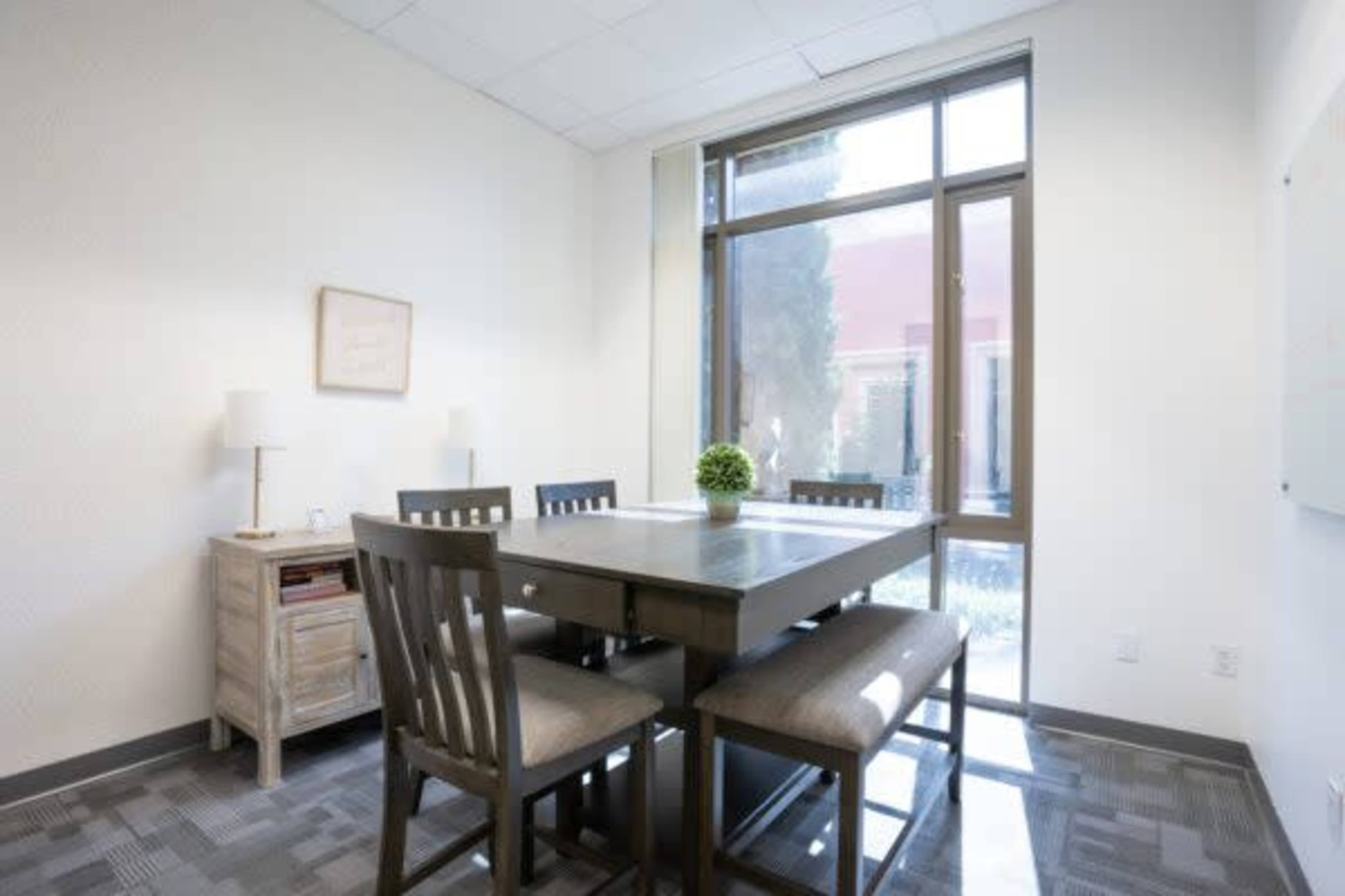A small conference room features a rectangular table surrounded by four chairs, with a lamp and a plant on a side table and a large window letting in natural light.