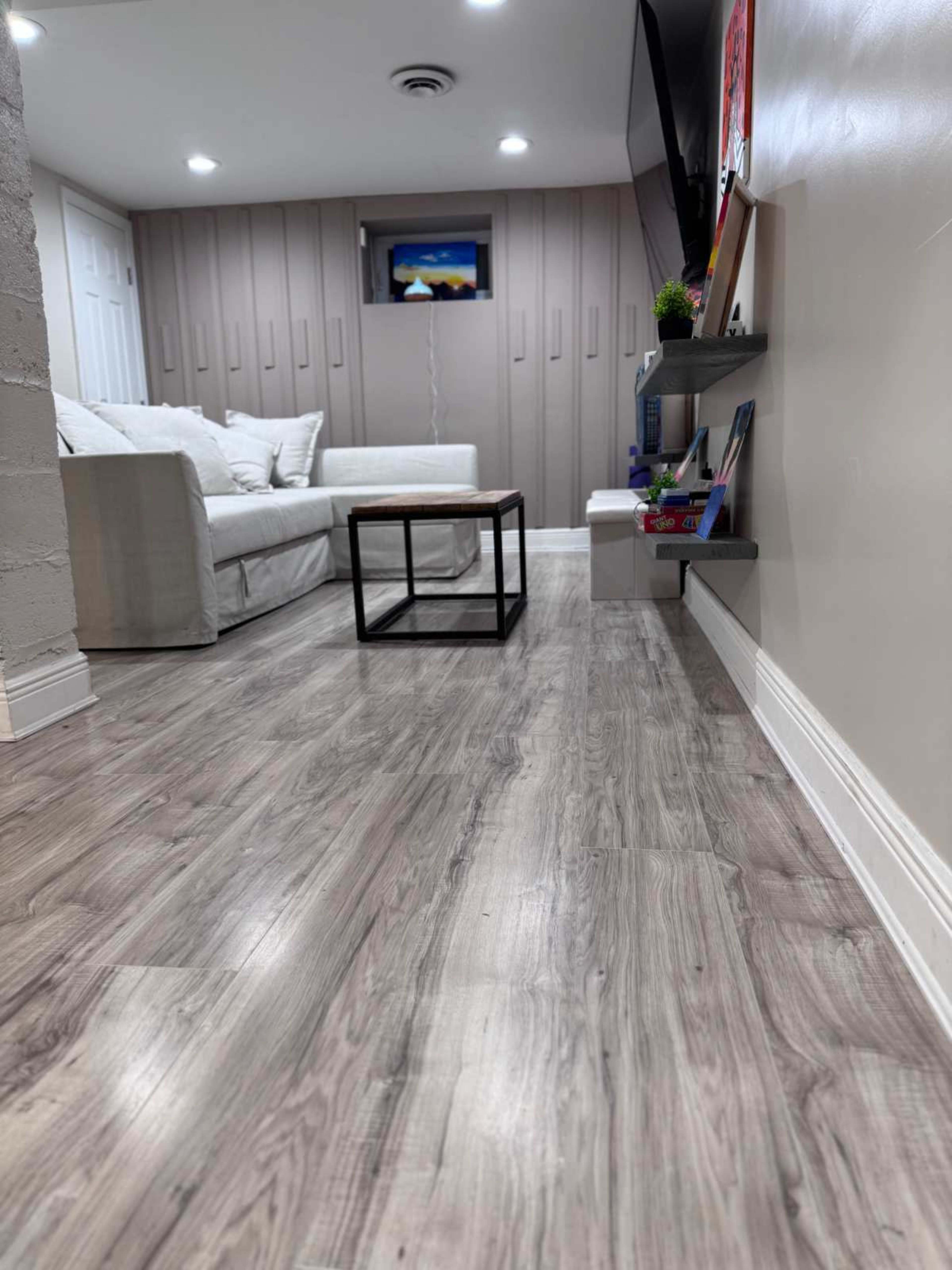 The image shows a cozy living room with a white sofa, a small coffee table, a television mounted on the wall, and shelves with books and decorative items.