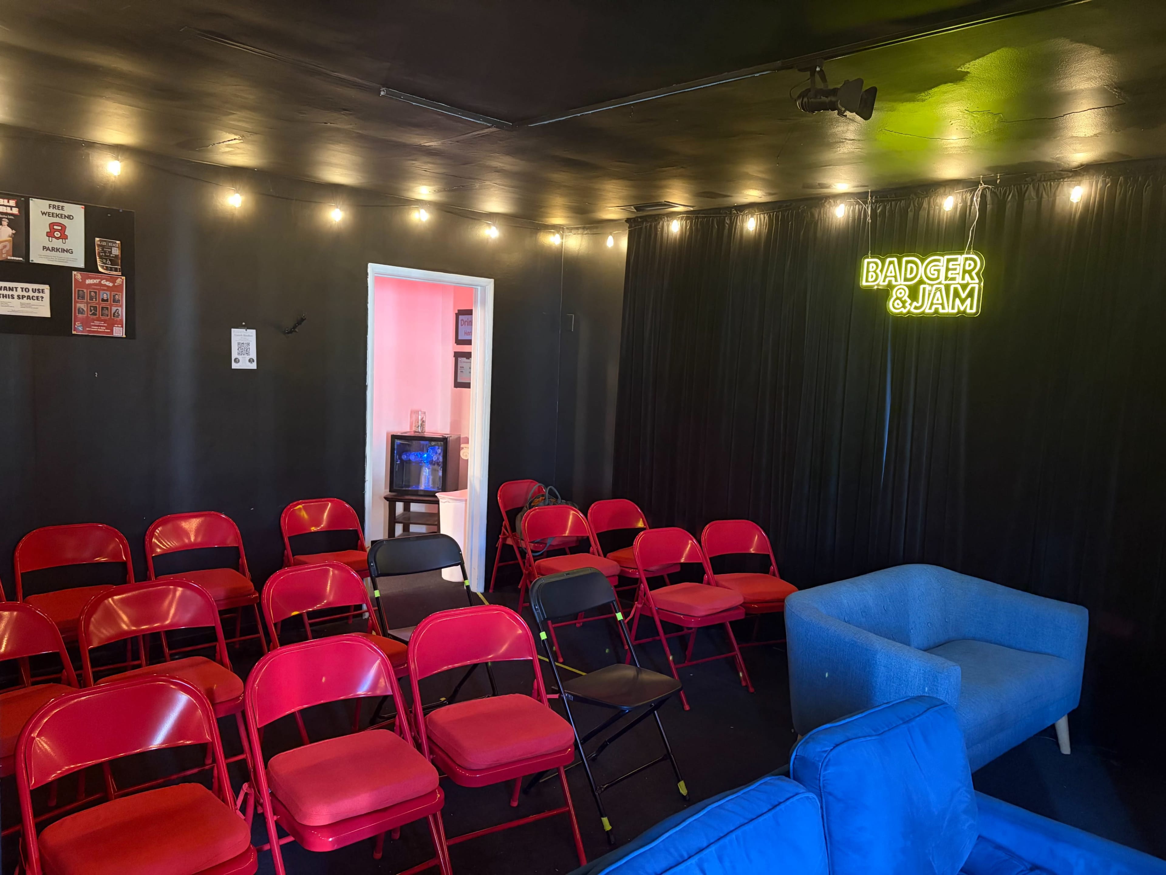 The image shows a small indoor venue featuring red folding chairs arranged in rows, a blue couch, and a neon sign that reads "BADGER & JAM."