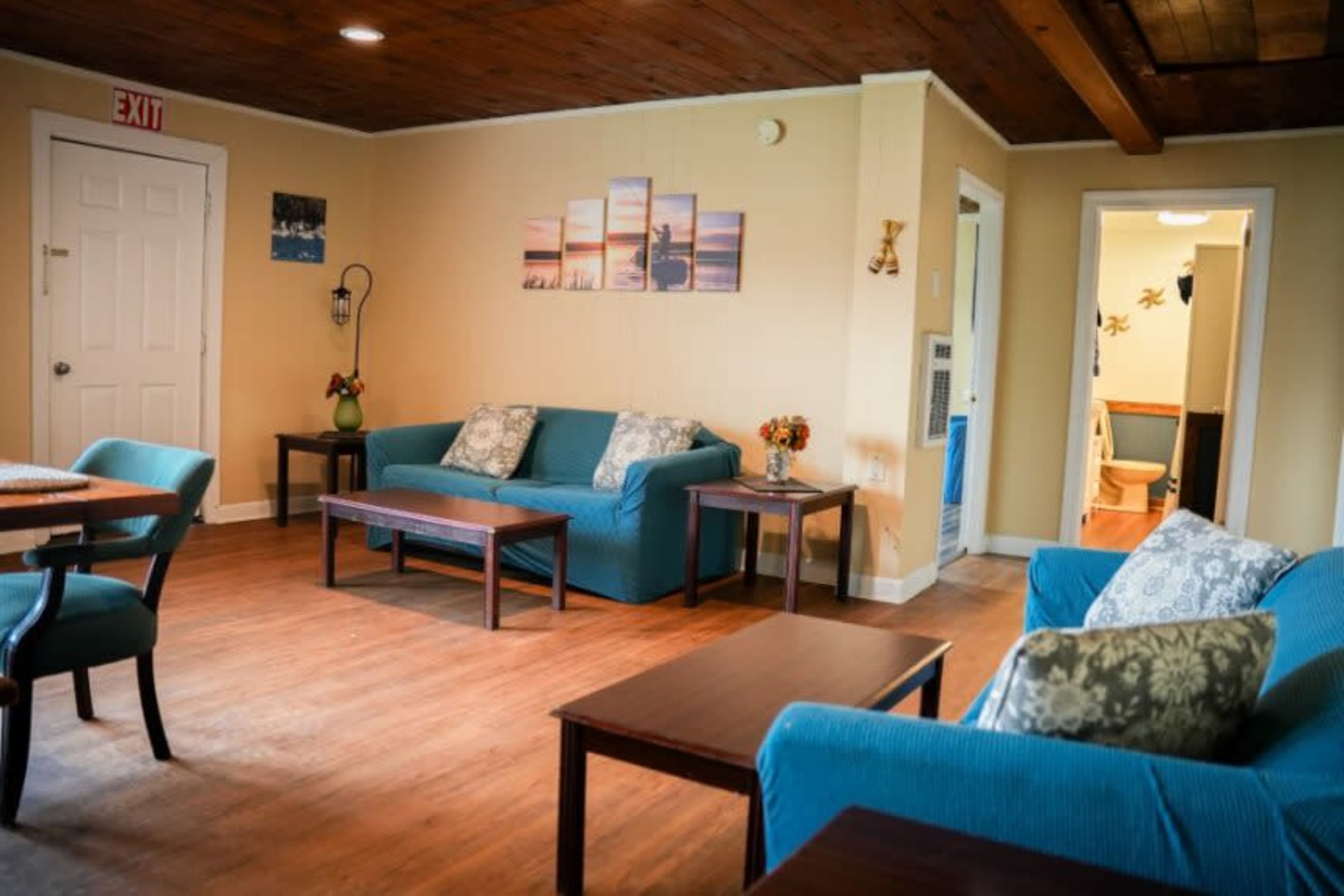 A living room with two blue sofas, wooden tables, and a doorway leading to a bathroom in the background.