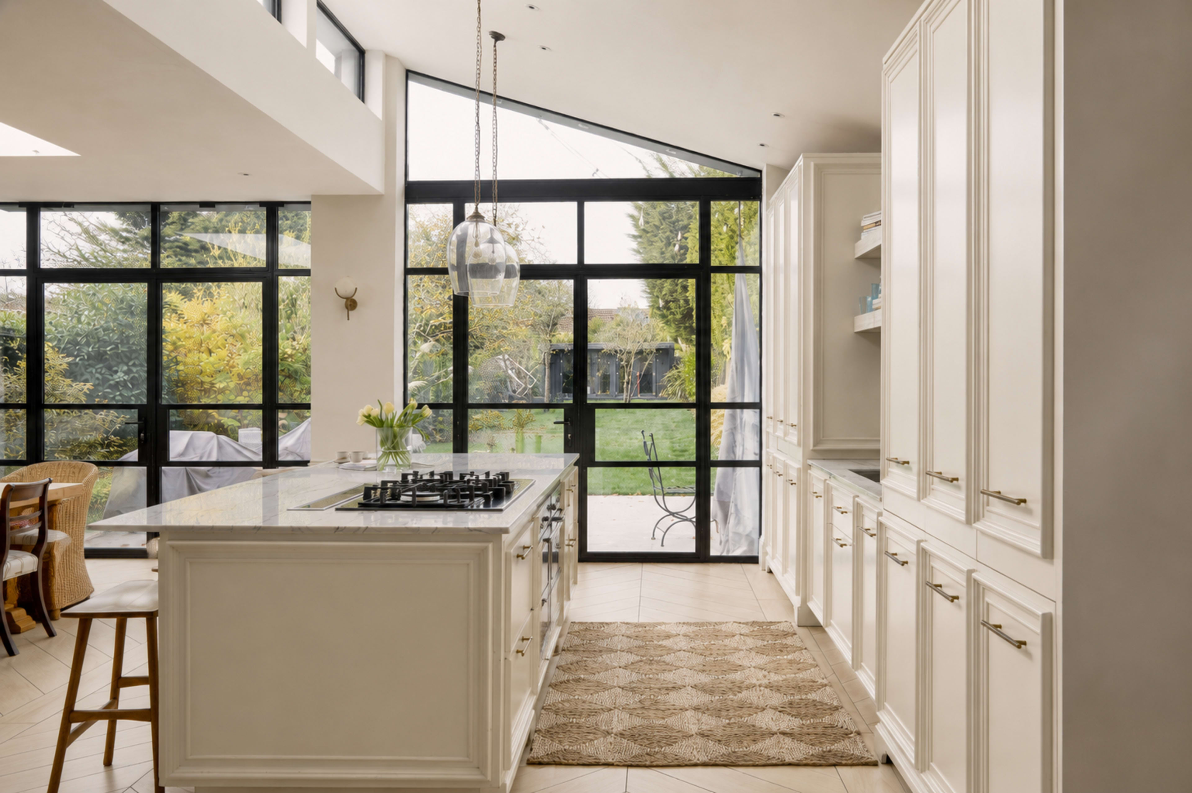 A spacious kitchen features a large island with a gas cooktop, white cabinetry, and expansive windows overlooking a green garden area.