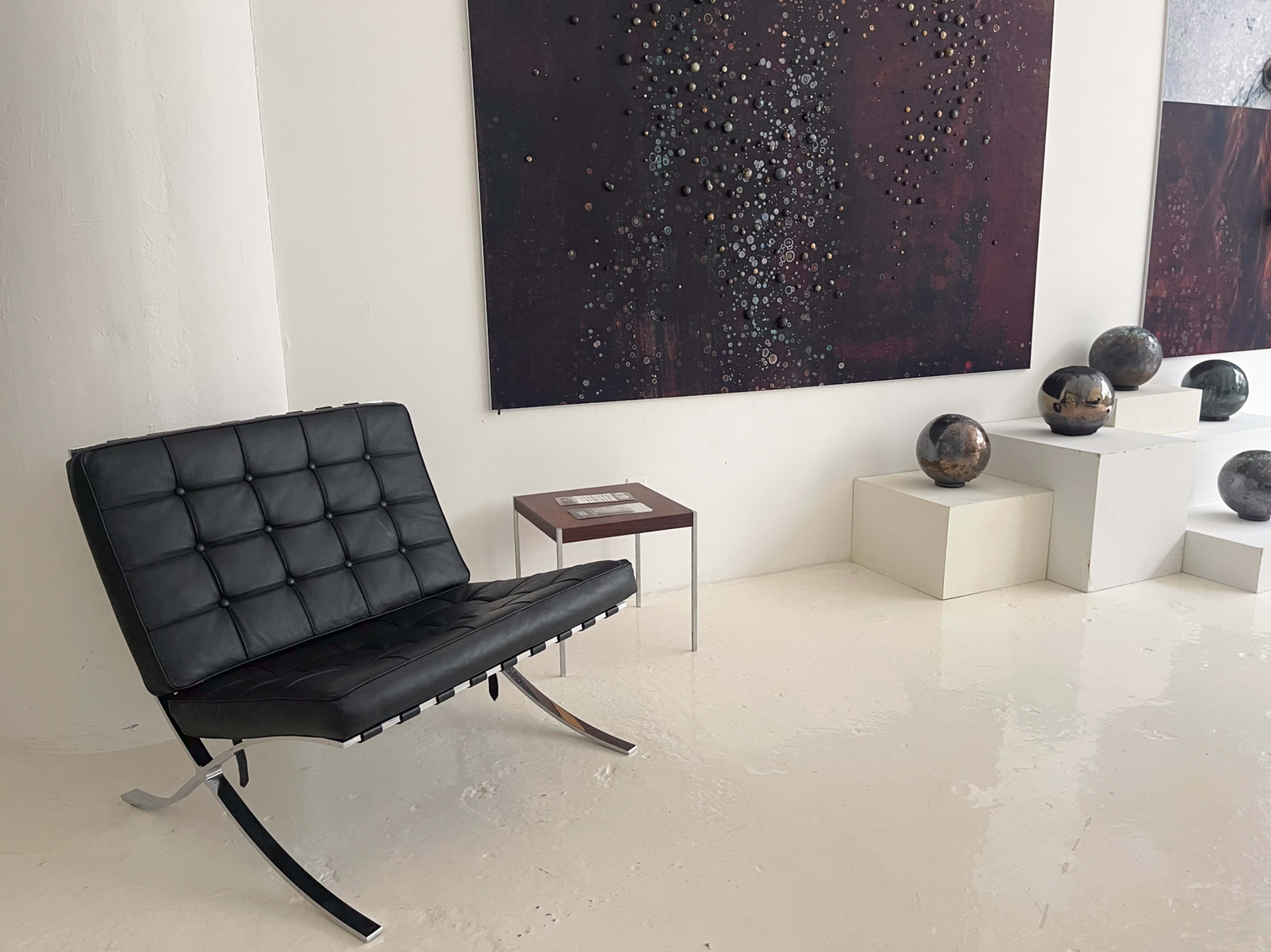 The image shows a modern black lounge chair beside a small square table, against a backdrop of large abstract art pieces and round decorative objects displayed on white pedestals.