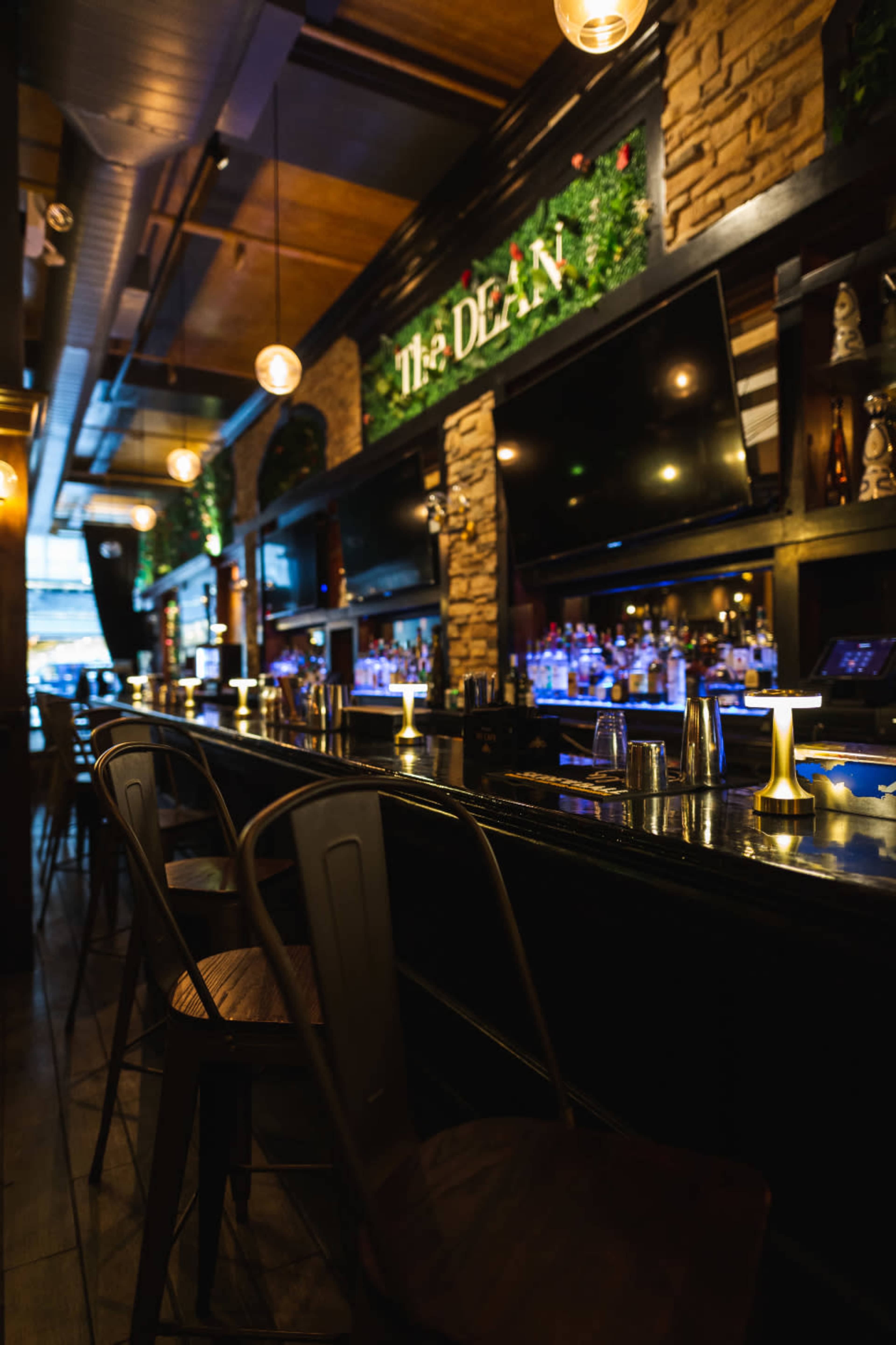 The image shows a dimly lit bar interior with a long countertop, a well-stocked liquor display, and decorative plants along the walls.