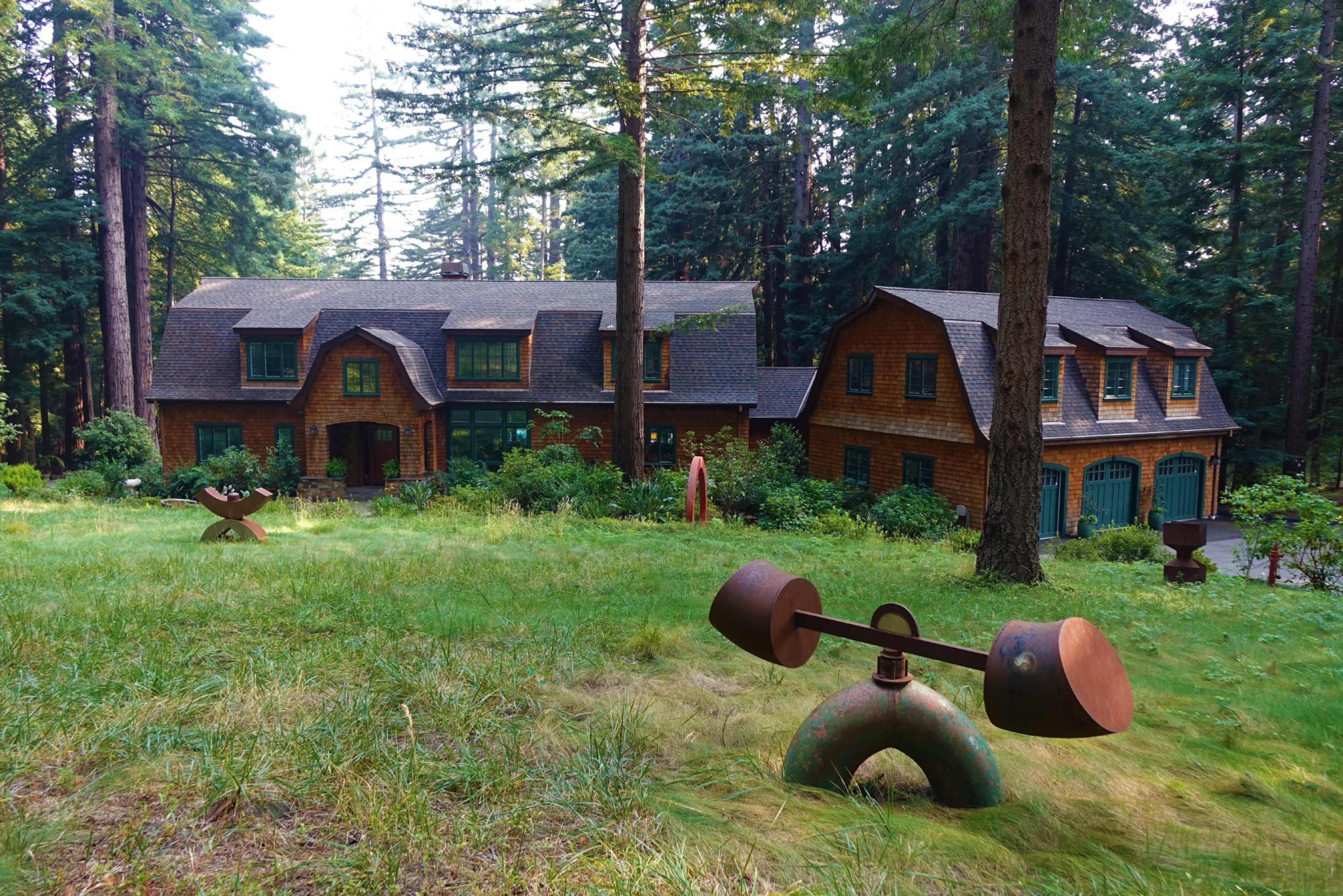 A large wood-paneled house is situated among tall trees, with several outdoor sculptures in the grassy yard.