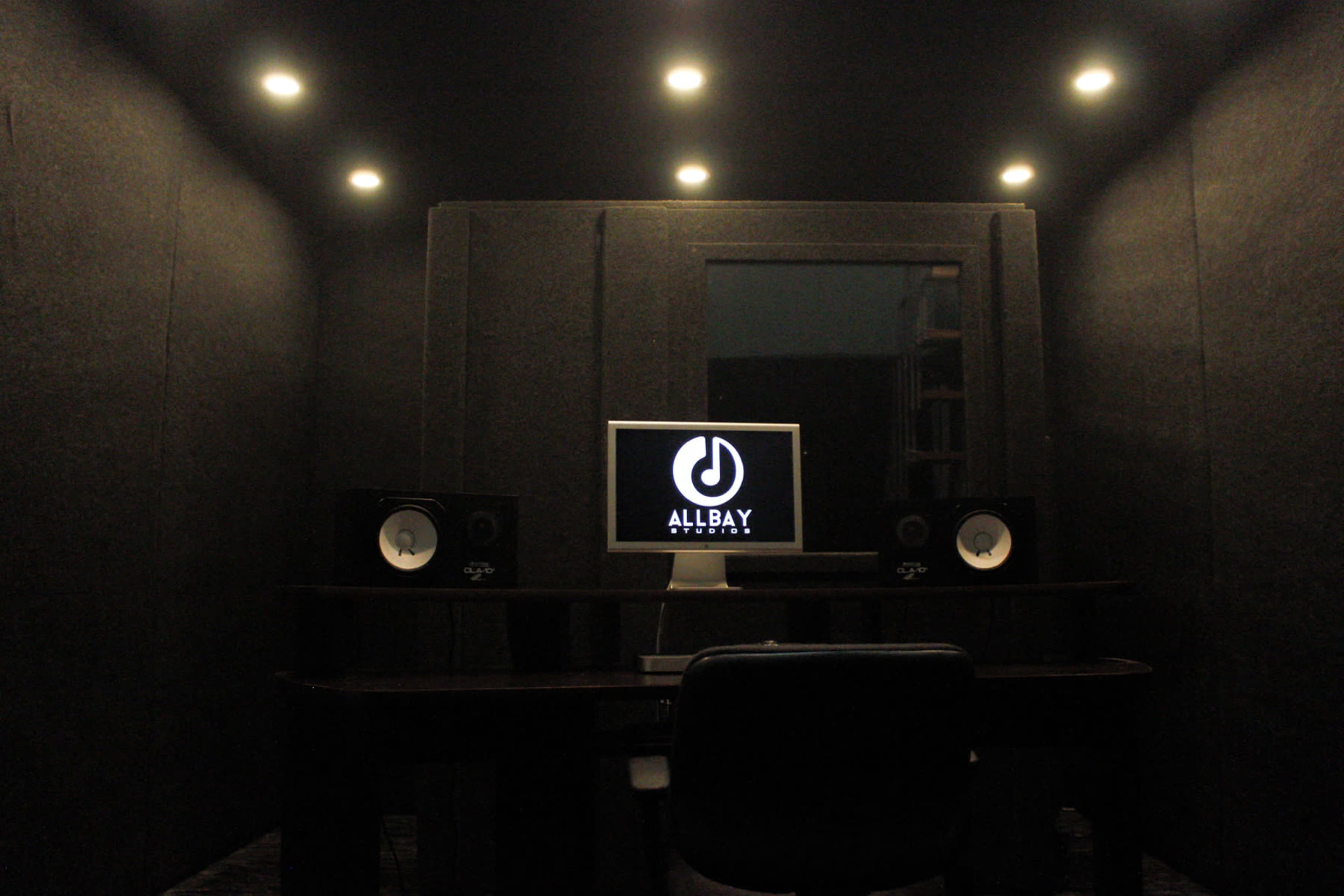 A soundproof recording studio features a computer monitor and speakers on a desk, with dark walls and ceiling lights.
