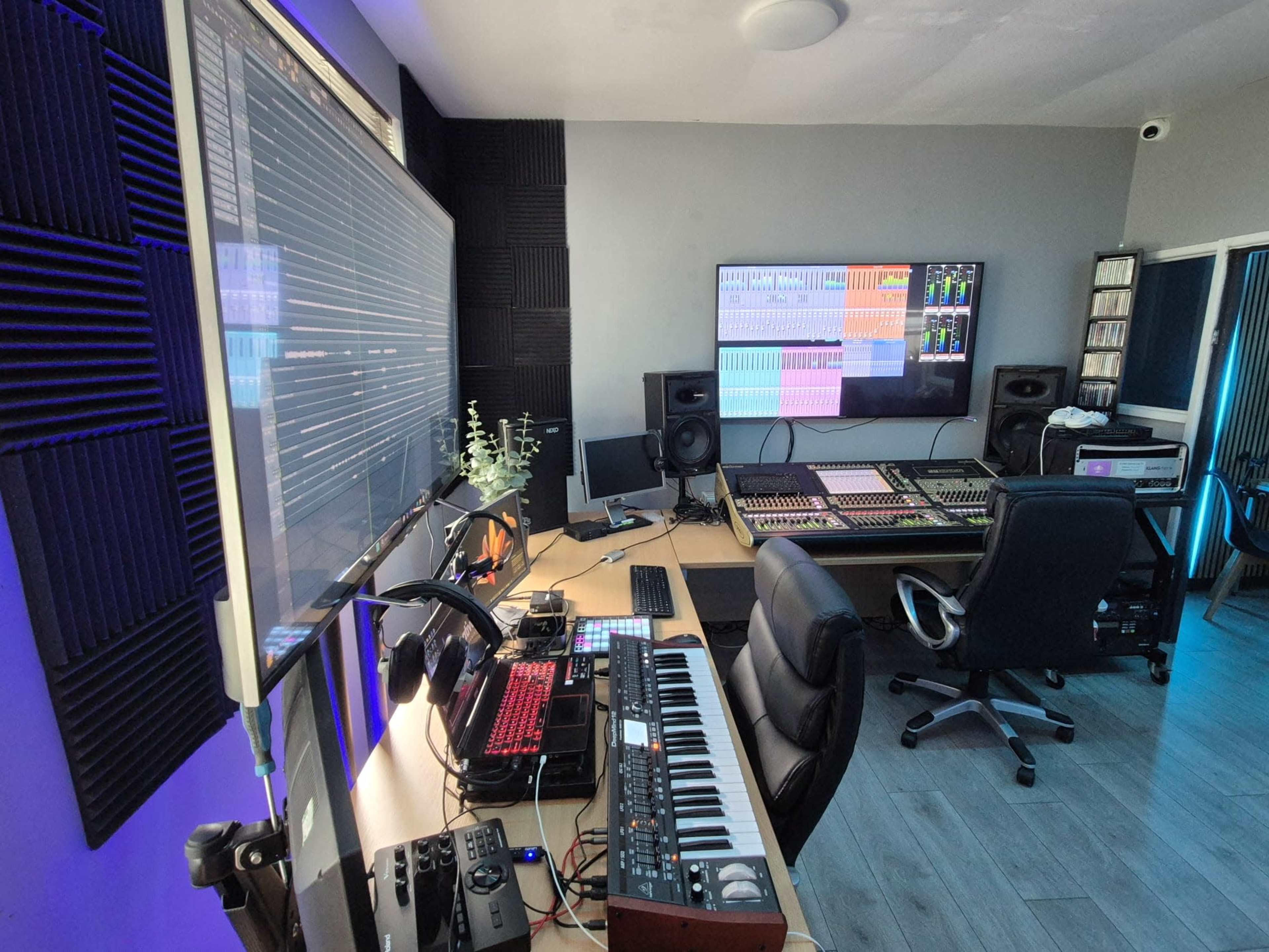 The image shows a modern music production studio featuring a large mixing console, computer monitors displaying audio software, and various instruments set up on a desk.