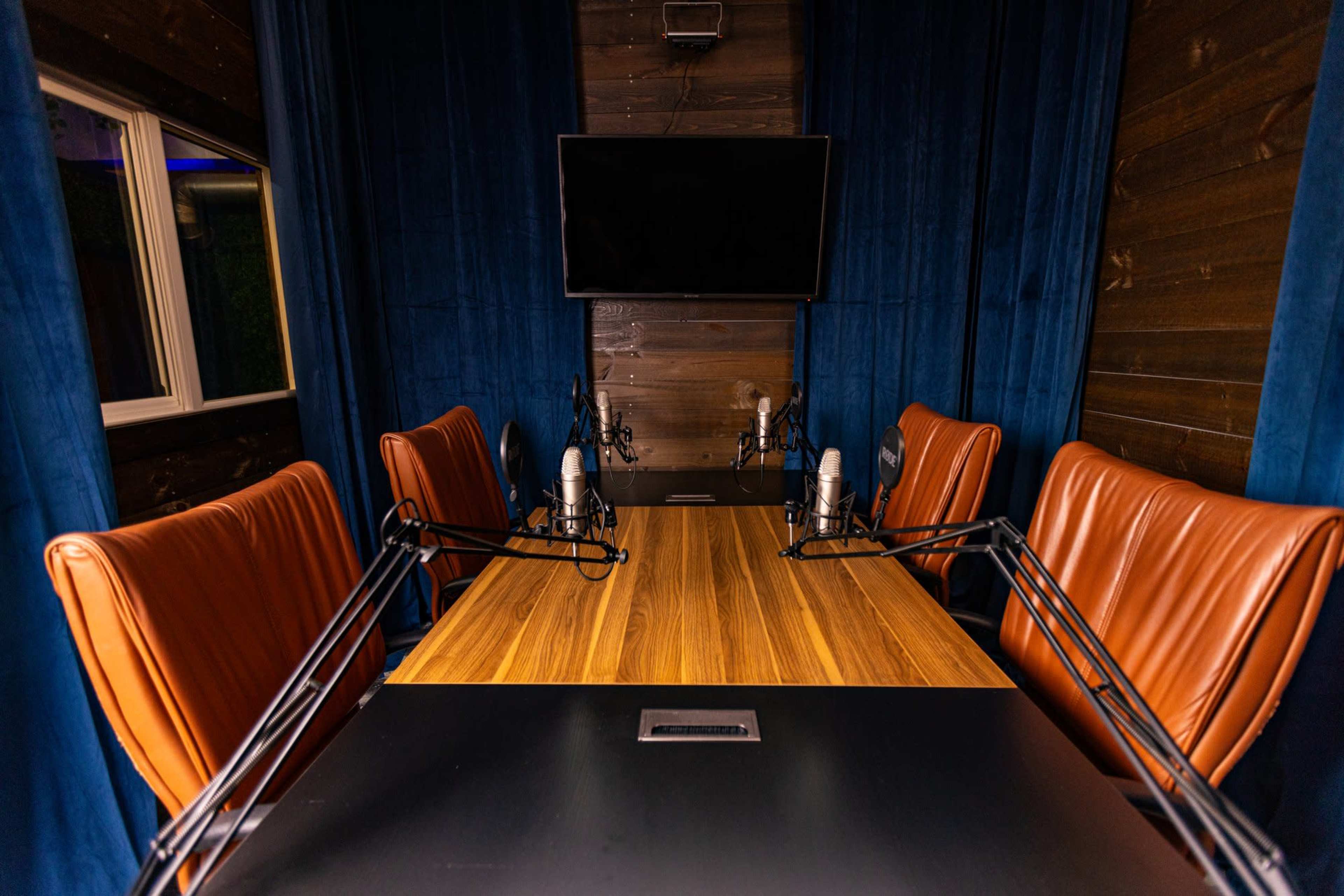 A conference room features a wooden table surrounded by four brown leather chairs and equipped with microphones, under a wall-mounted television.