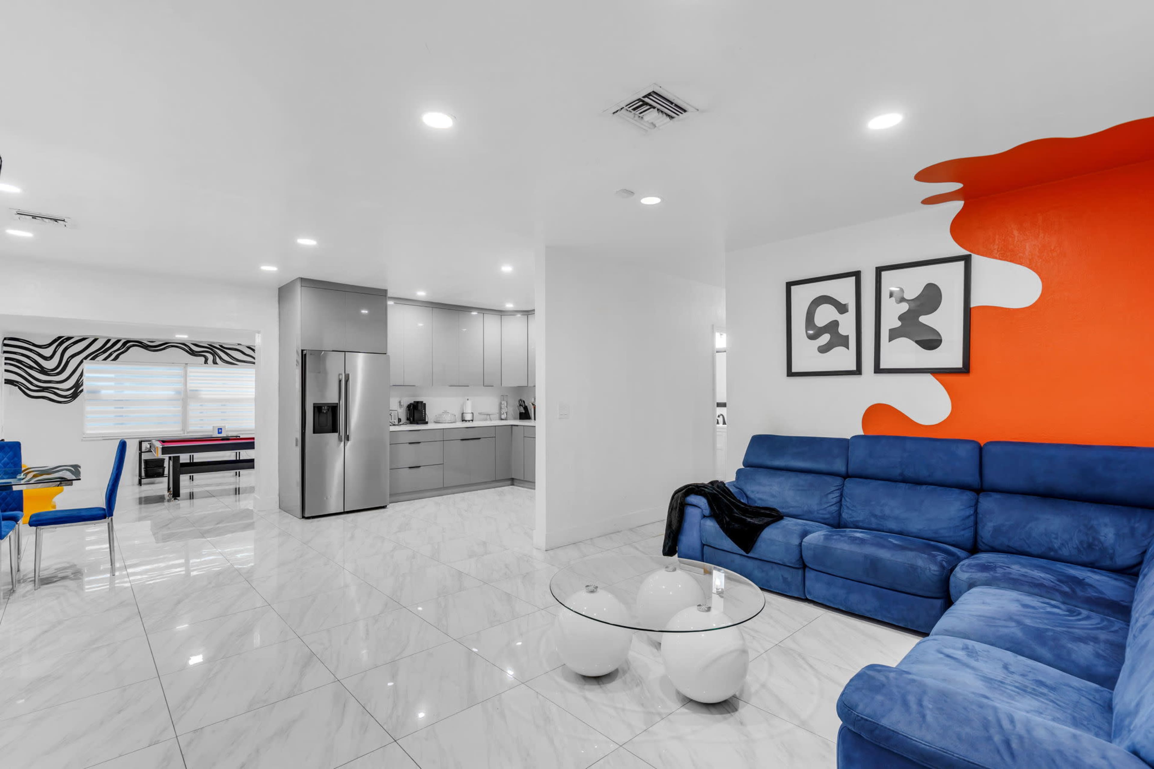 The image shows a modern living space featuring a blue sectional sofa, a glass coffee table, and a kitchen area with sleek cabinetry and stainless steel appliances.