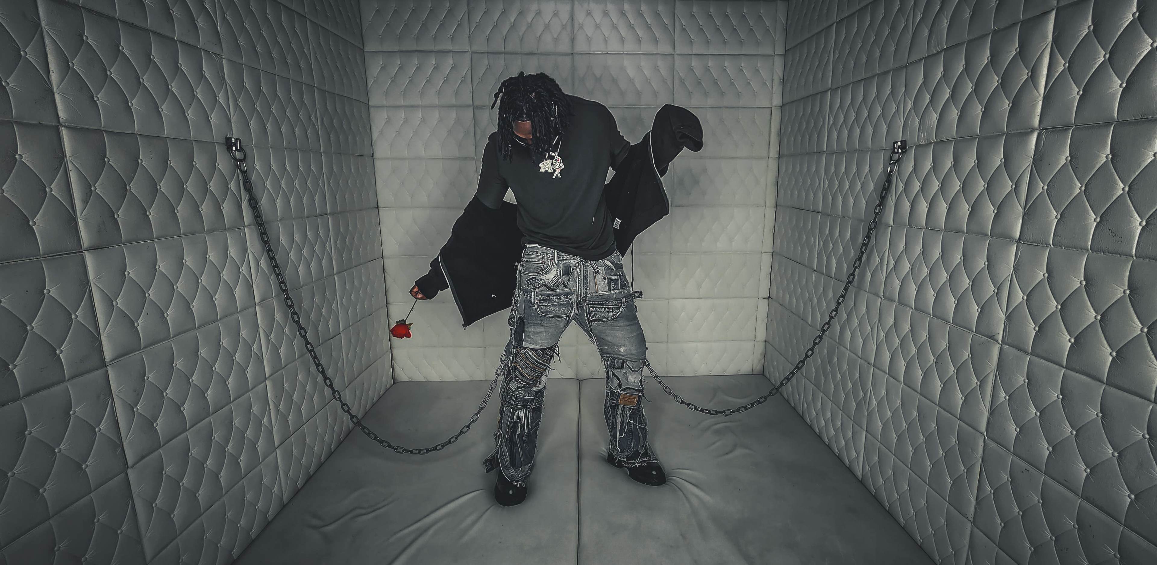 A person is standing in a small, enclosed space with padded walls, holding a single red rose while wearing a black outfit and distressed jeans.