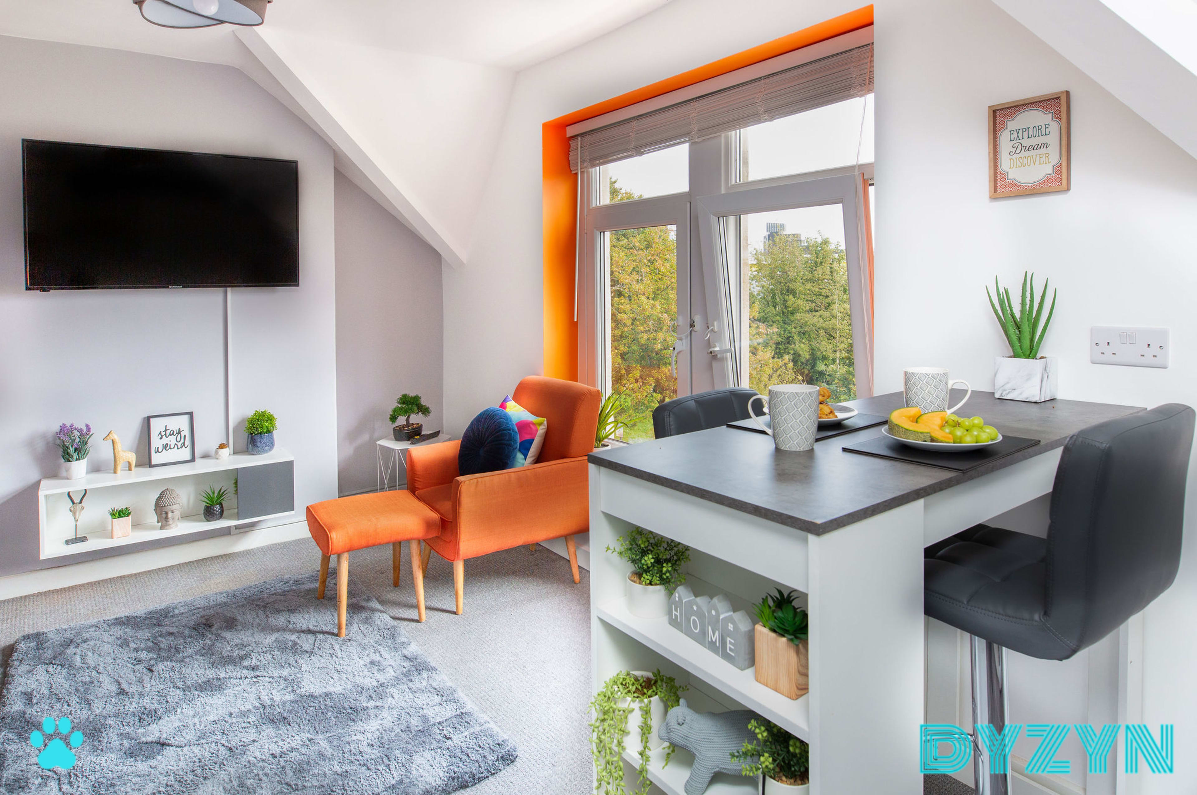 A modern living space features an orange-accented chair, a gray rug, a small dining table set with fruit, and large windows showcasing greenery outside.