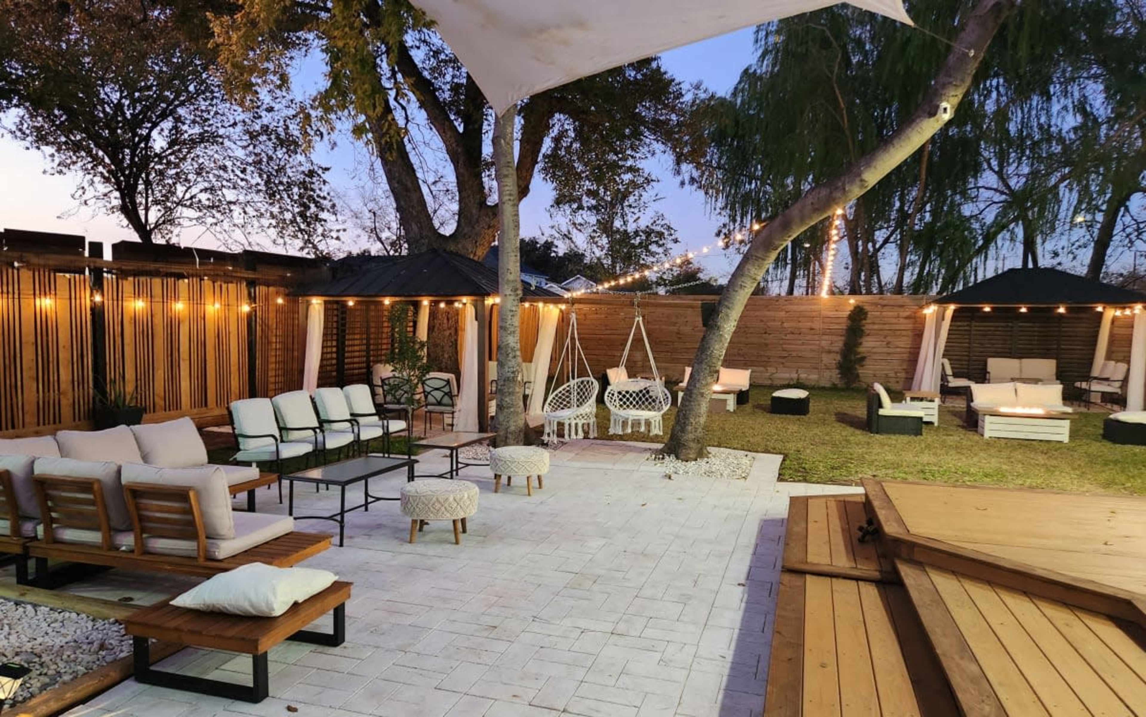 The outdoor lounge area features seating arrangements, decorative lights, and a grassy space surrounded by trees and wooden fencing.