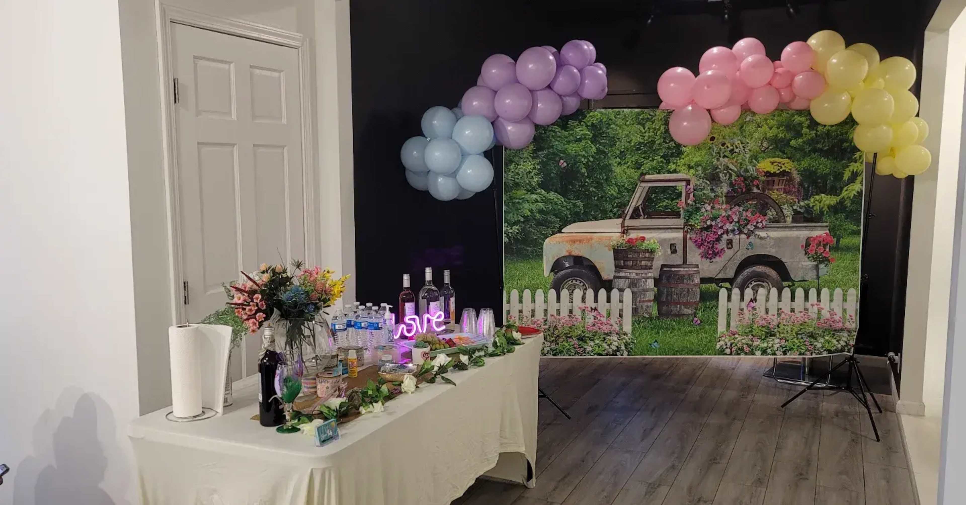 A table with floral arrangements and drinks is set up in front of a backdrop featuring an antique truck surrounded by greenery and a white picket fence, adorned with a colorful balloon arch.