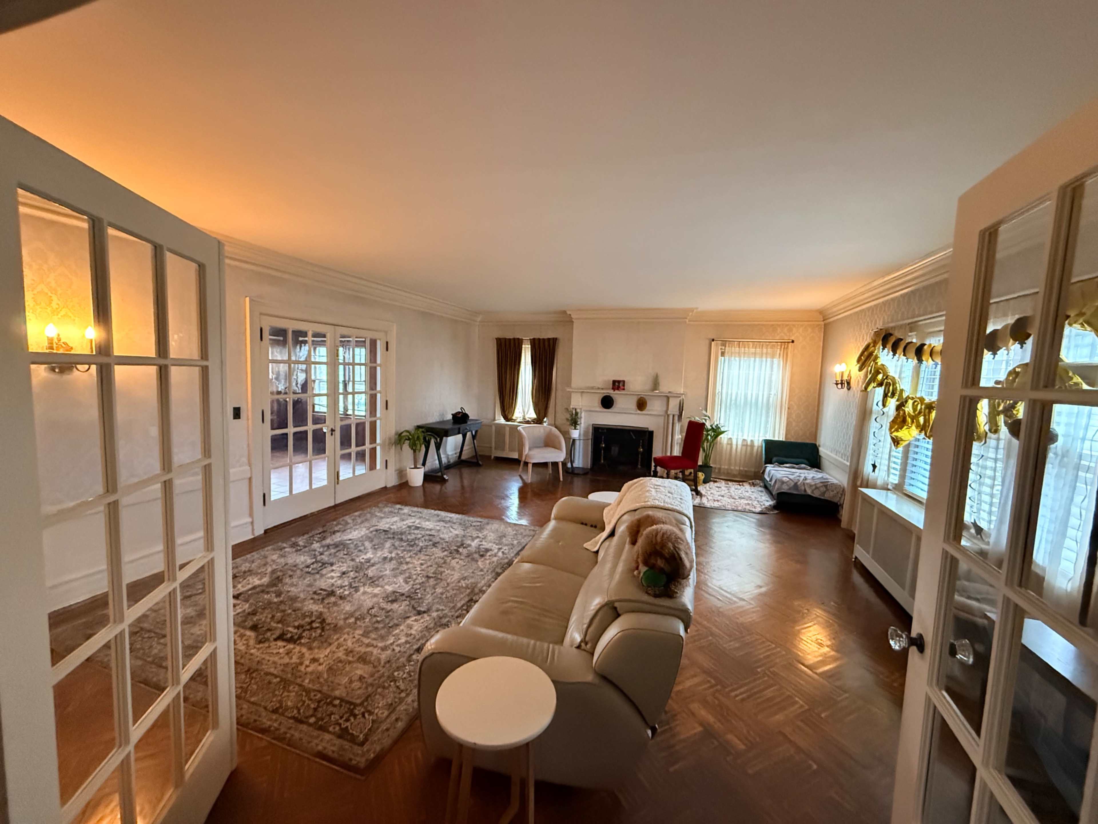 The room features a spacious layout with a beige sofa, a round white table, and large windows adorned with curtains, creating a bright atmosphere.