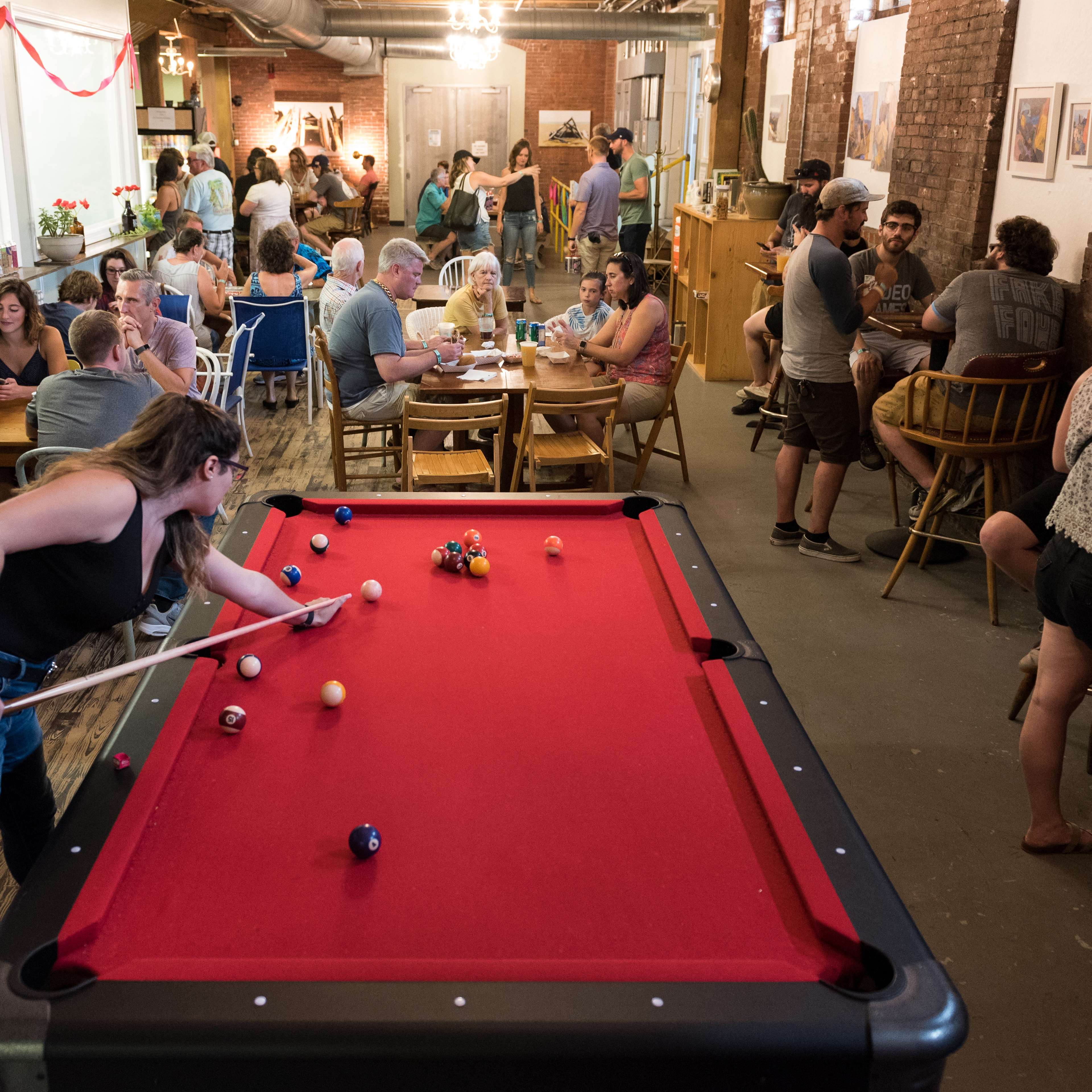 A crowded social space features people engaging in various activities, including playing pool, dining, and conversing.