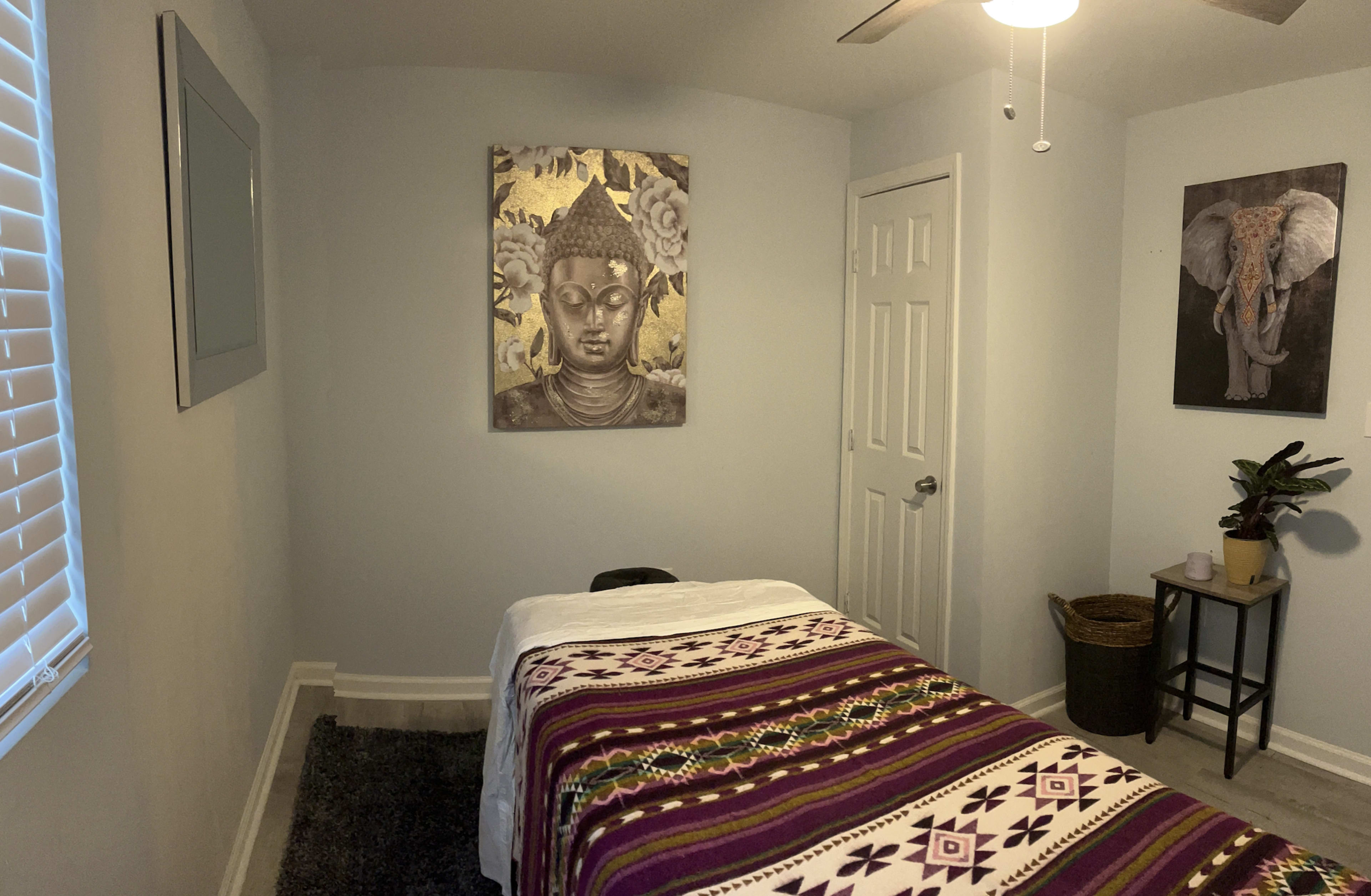 The image shows a serene room with a massage table covered by a patterned blanket, a Buddha artwork on the wall, and an elephant picture beside a small plant and a woven basket.