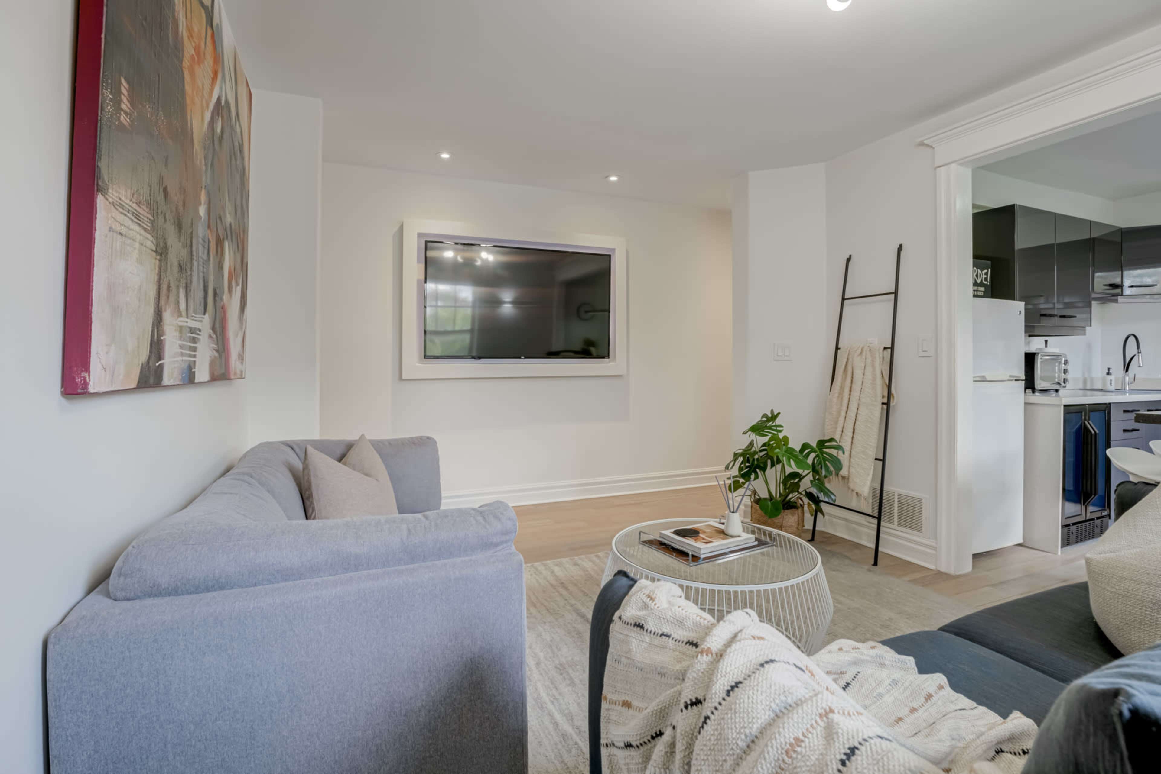 A modern living room features a gray sofa, a round coffee table, and a wall-mounted TV, with an open view toward a kitchen area.