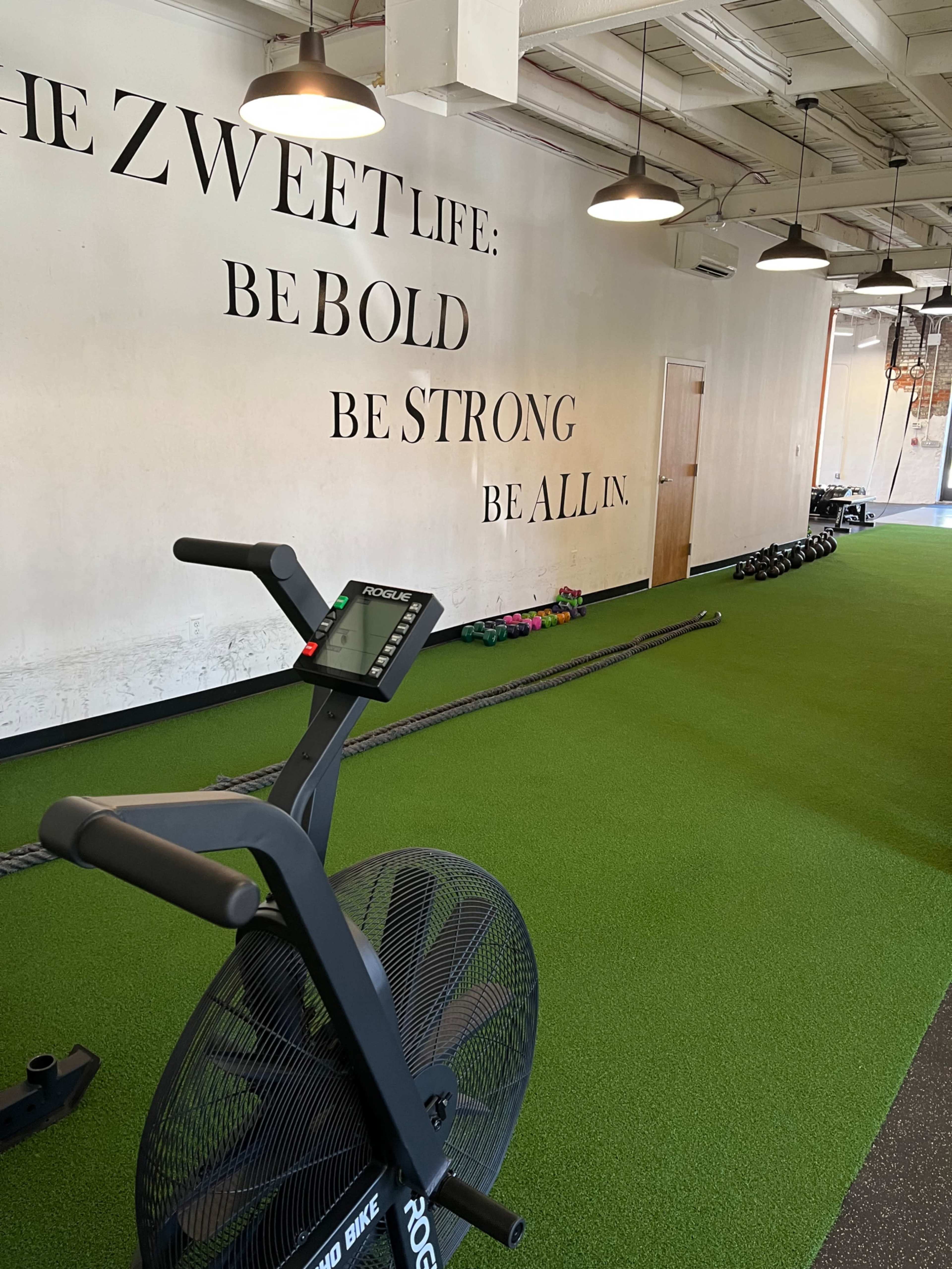The image shows a modern gym with a stationary bike in the foreground, green turf flooring, and motivational text on the wall.