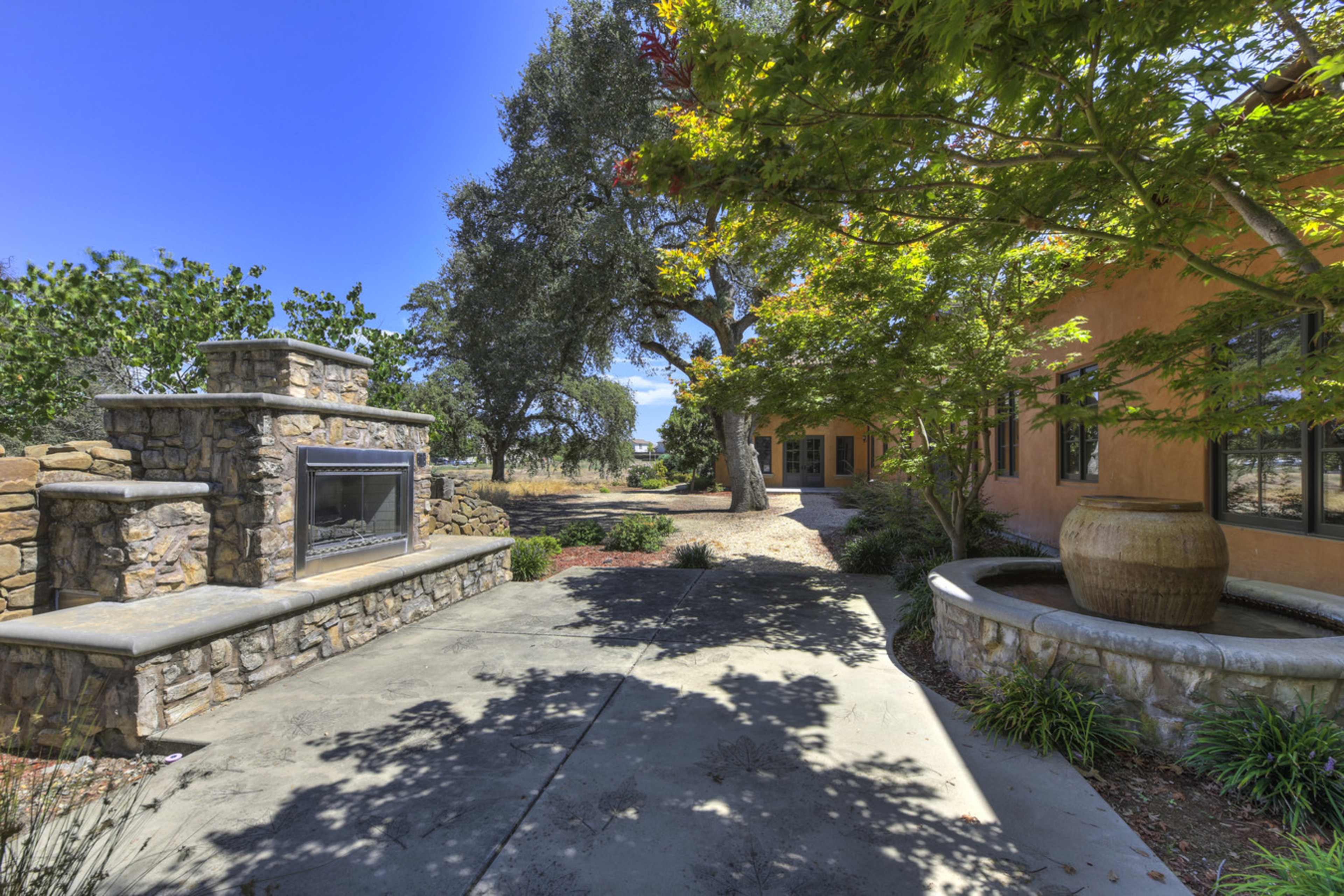 A stone outdoor fireplace next to a pathway lined with trees and a decorative water feature beside a stucco building.
