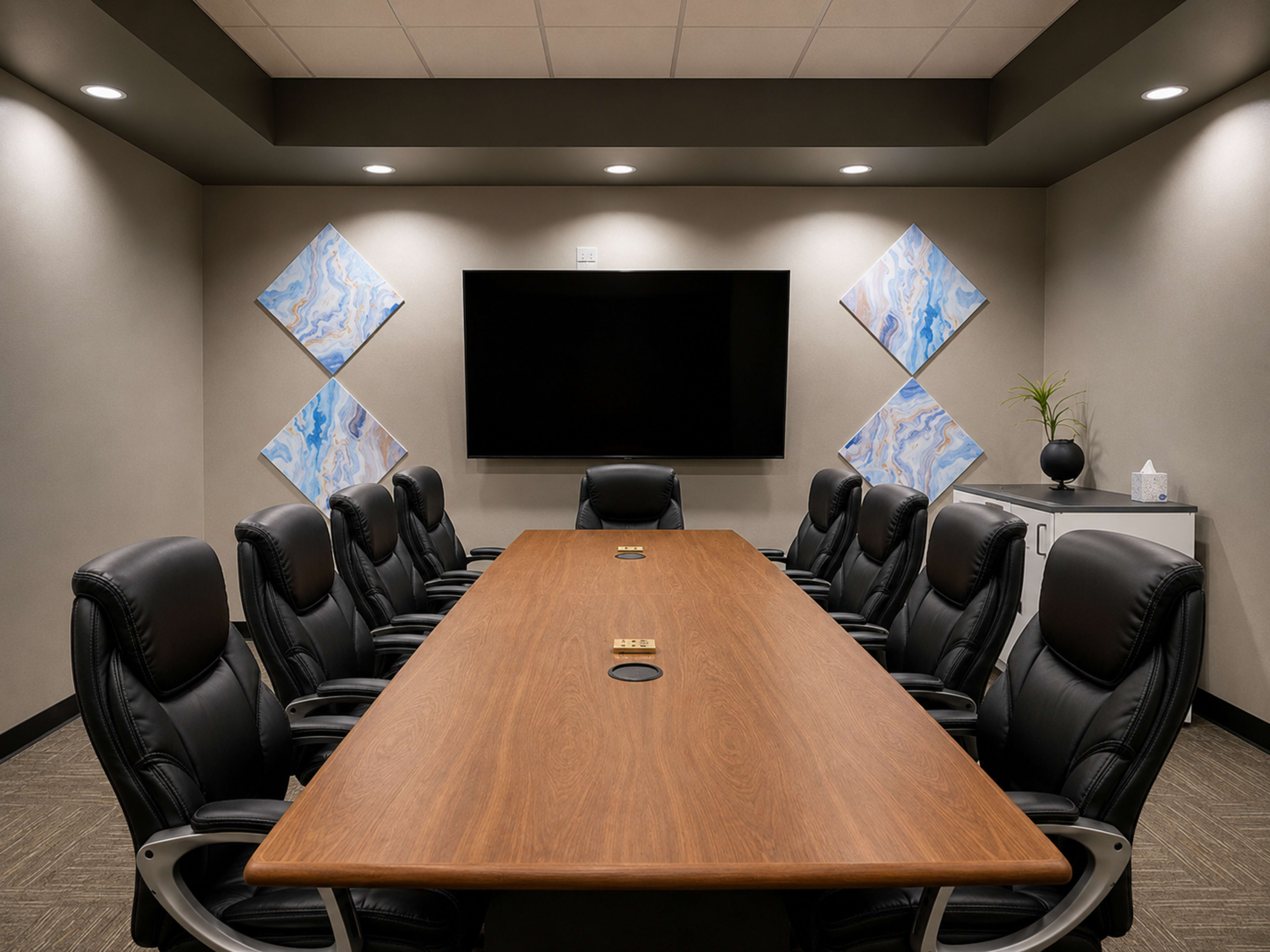 A modern conference room features a long wooden table surrounded by eight black ergonomic chairs, a large screen mounted on the wall, and abstract art pieces on adjacent walls.