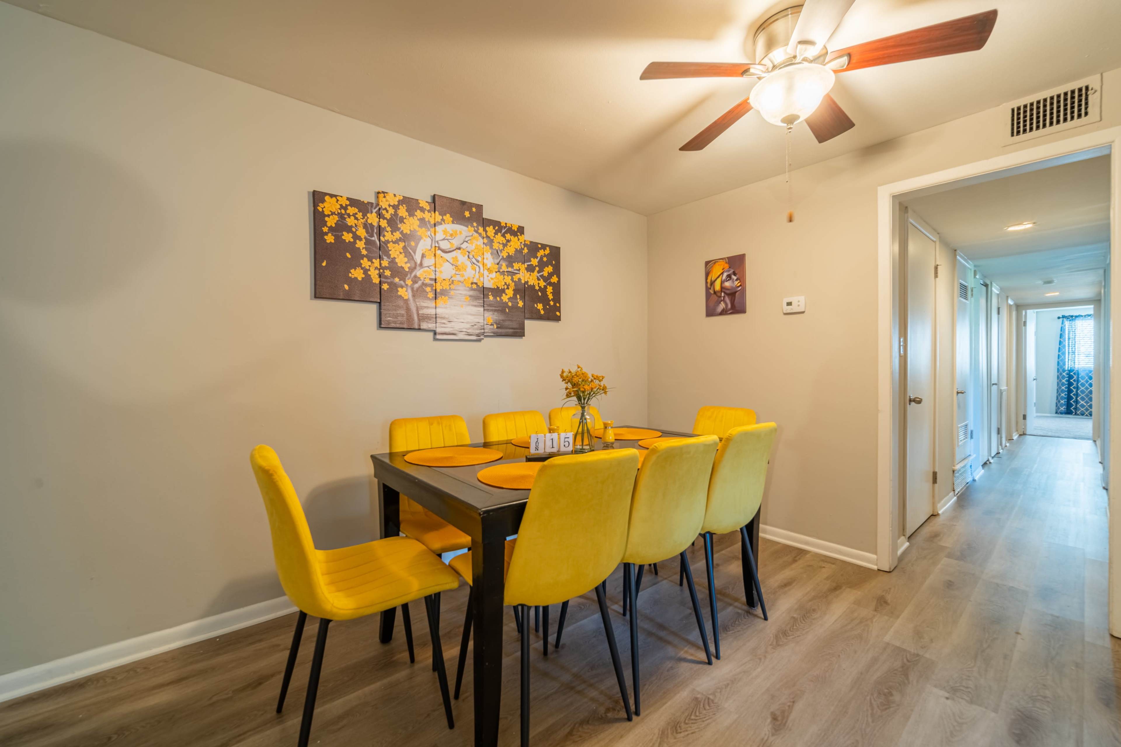 A dining area features a black table surrounded by yellow chairs, with artwork of a sunset and a vase of flowers on the table.
