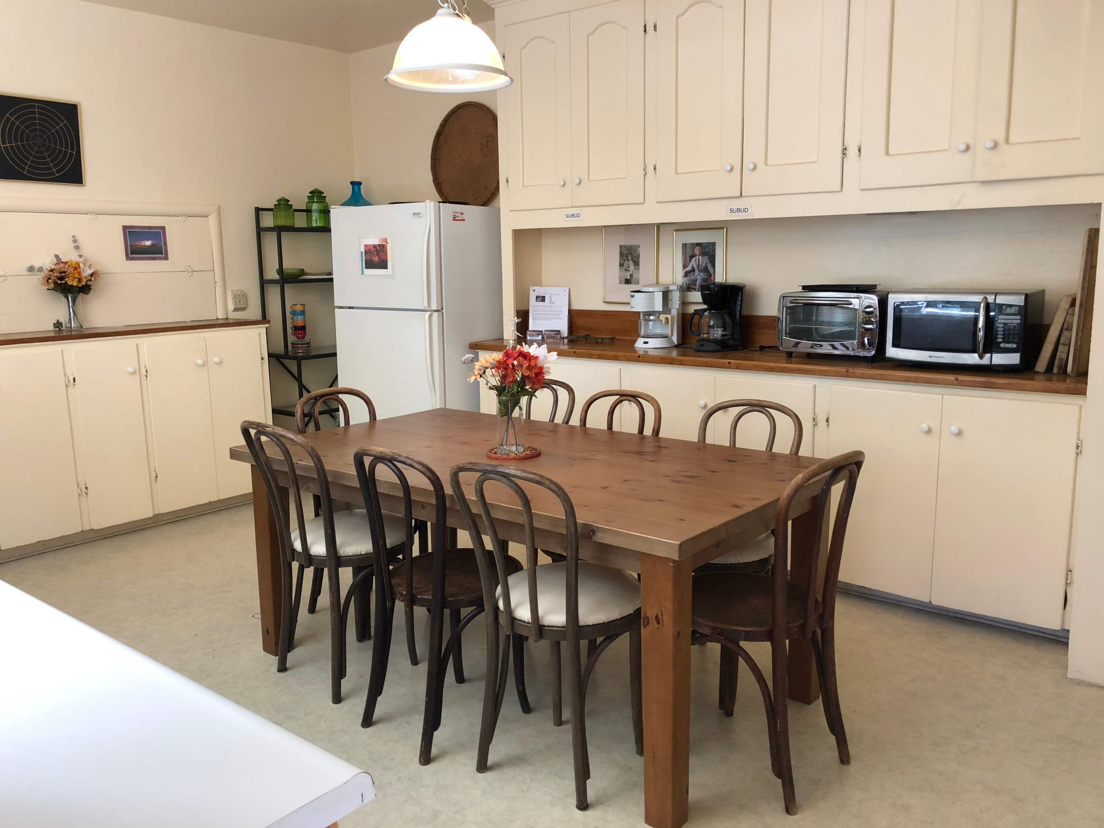A simple kitchen with cream-colored cabinets, a wooden dining table surrounded by chairs, and various appliances including a refrigerator and microwave.