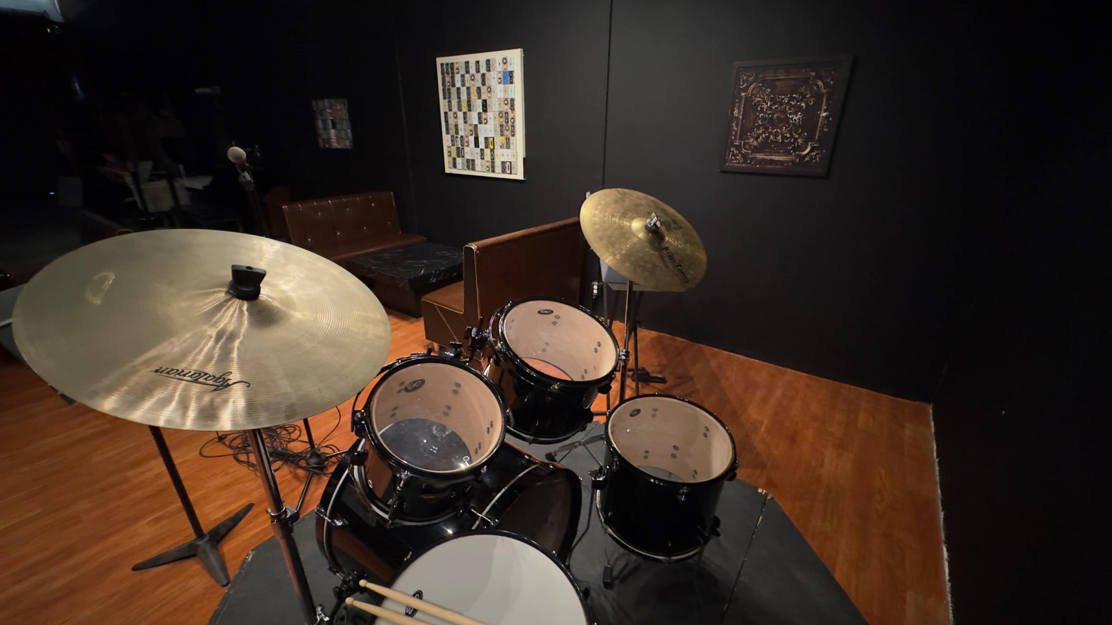 A drum set is positioned on a raised platform in a dimly lit room featuring dark walls and a few pieces of wall art.