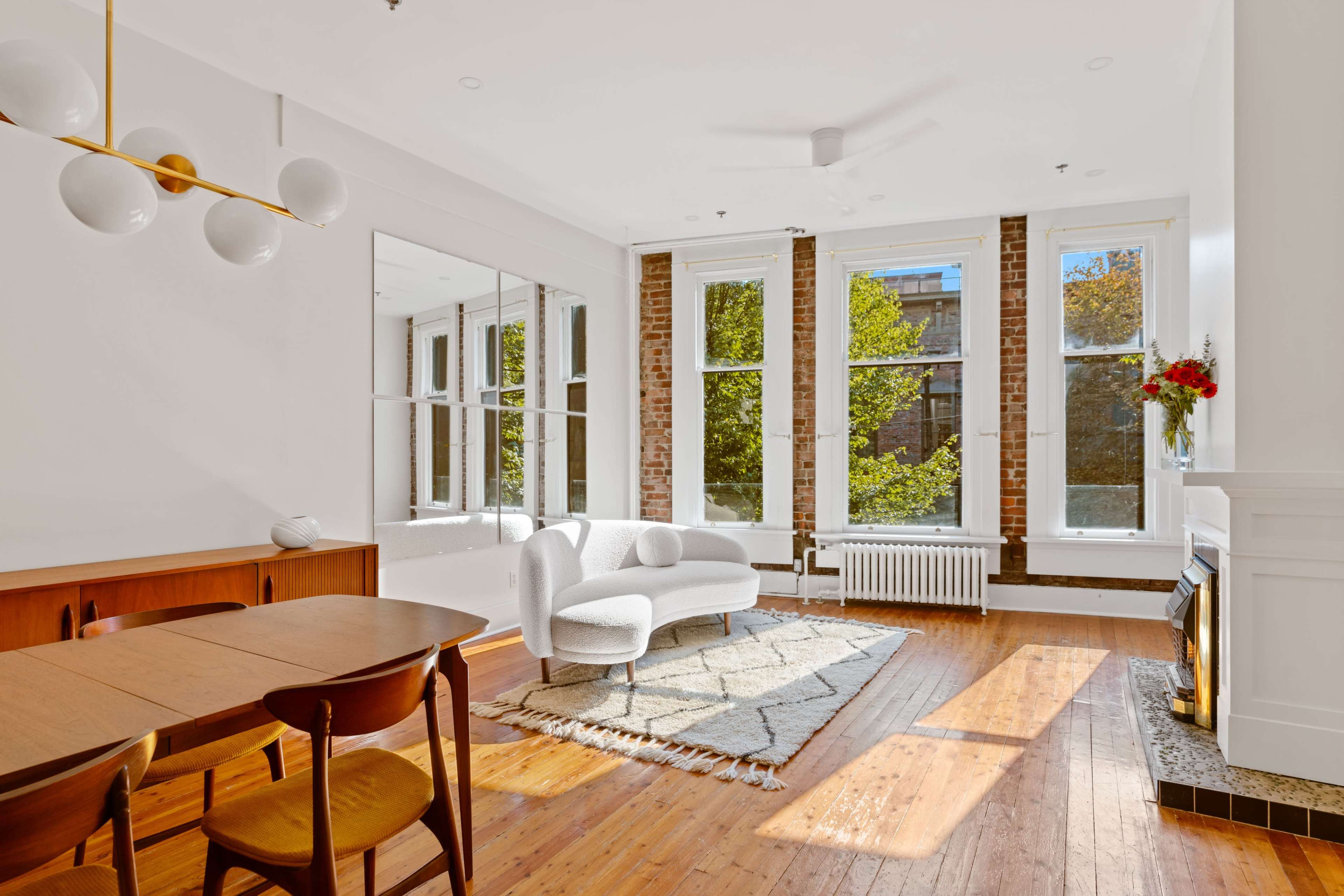 A bright living space features large windows, a white sofa, a wooden dining table, and hardwood flooring.