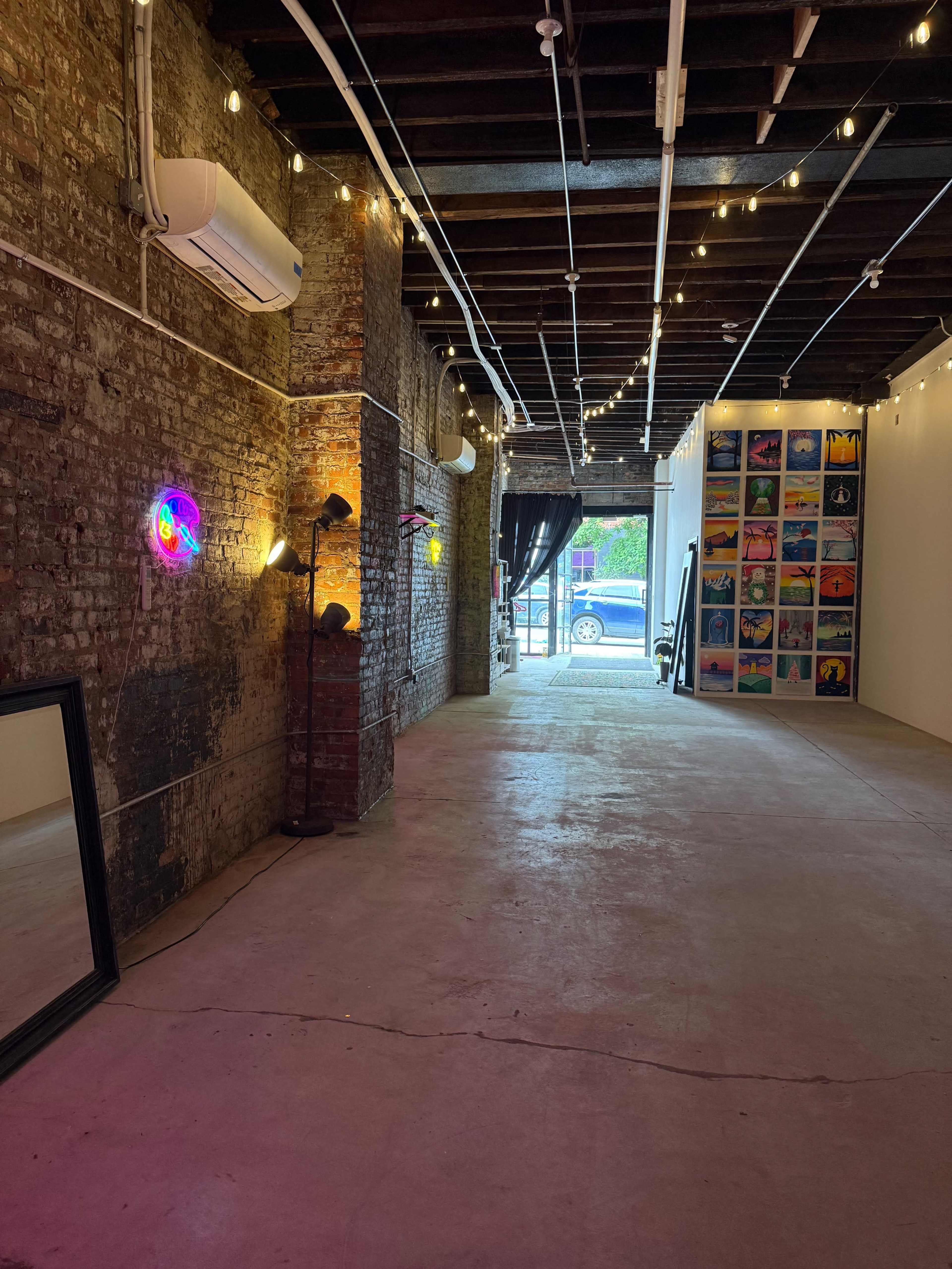 The image shows a spacious art gallery with exposed brick walls, string lights overhead, and colorful artwork displayed on one wall.