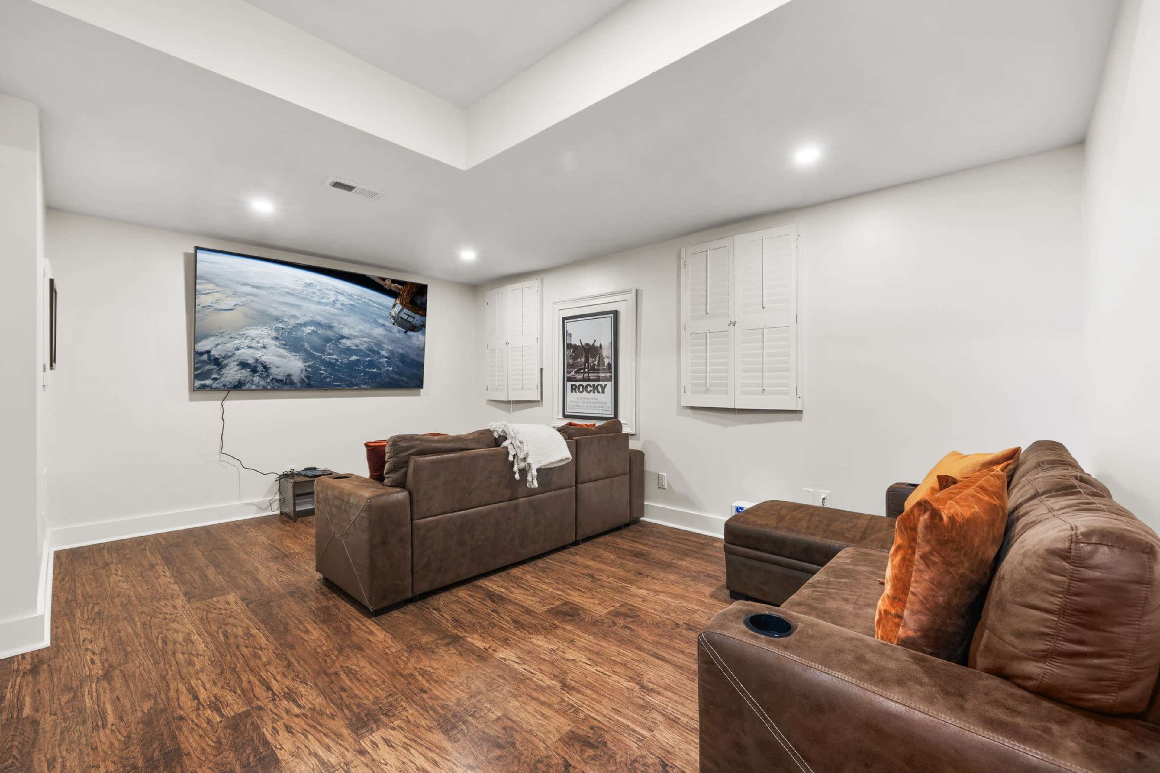 The image shows a modern basement living room with a large wall-mounted screen displaying a space scene, two brown sofas, and hardwood flooring.