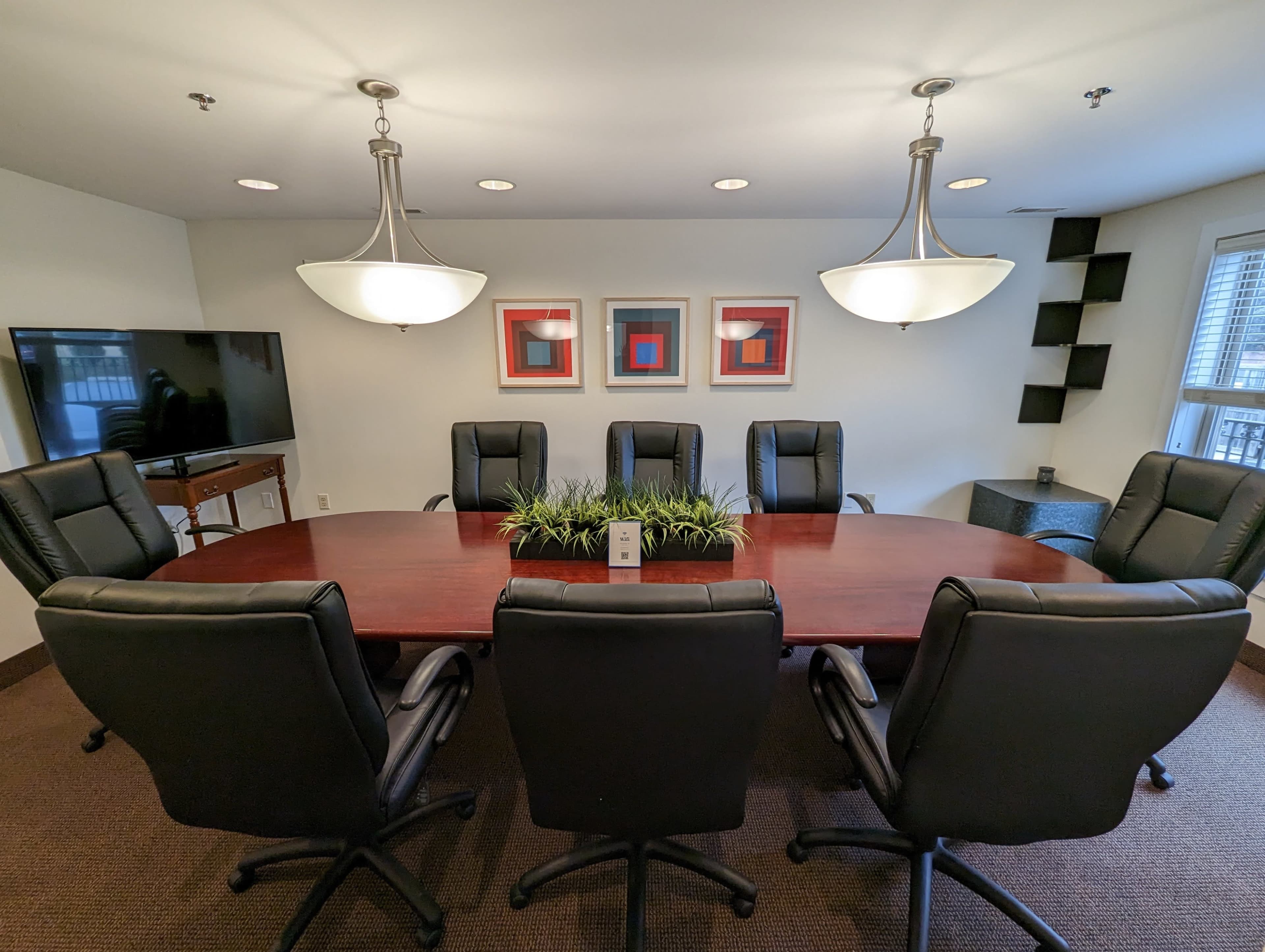 A modern conference room features a large wooden table surrounded by eight black leather chairs, with a decorative plant centerpiece and two pendant lights overhead.