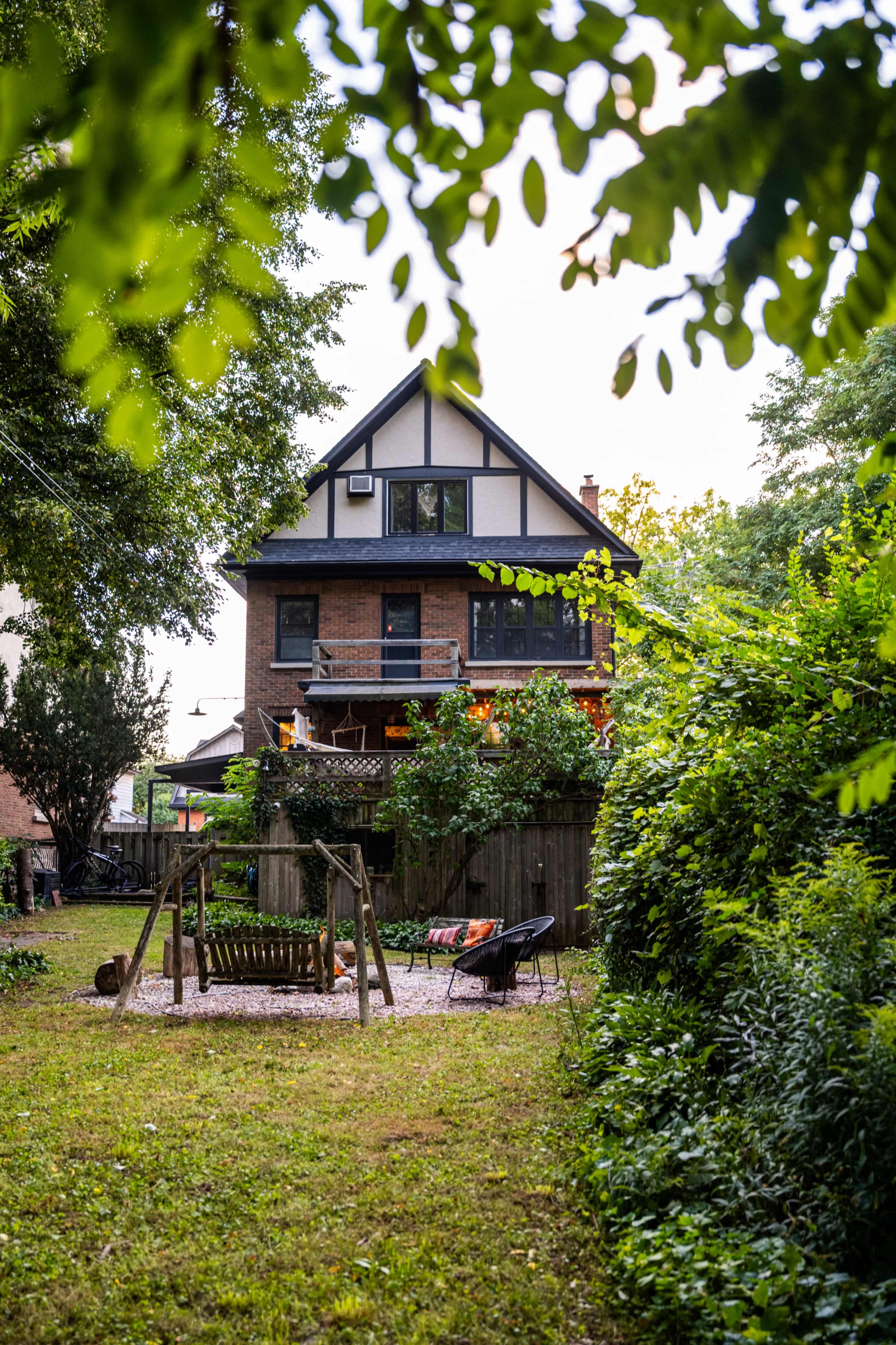 A two-story house with a balcony and a backyard featuring a swing set is surrounded by trees and greenery.