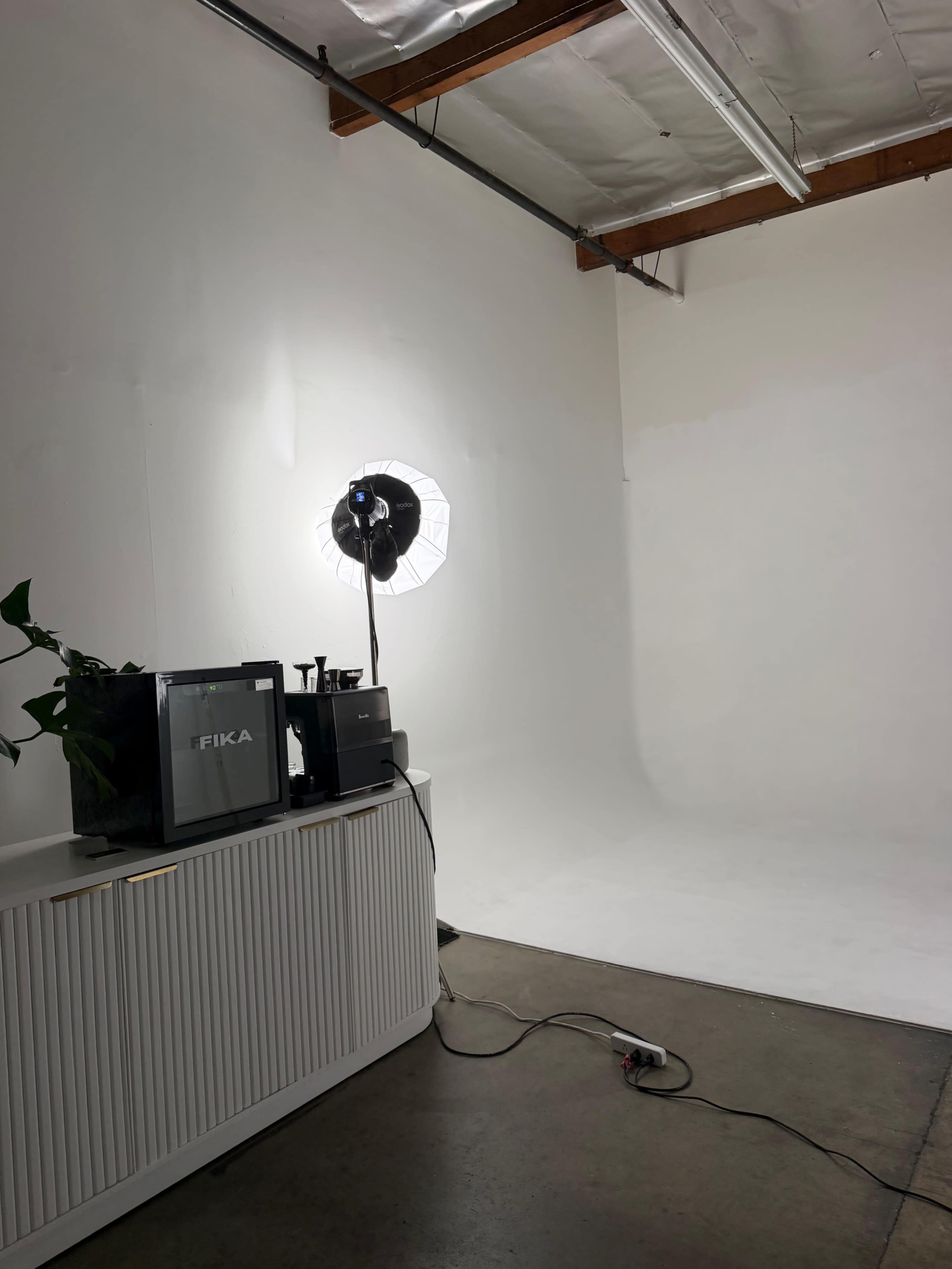 The image shows a minimalistic photography studio with a light source, a television displaying the word "FIKA," and a potted plant on a sleek white cabinet against a plain white backdrop.