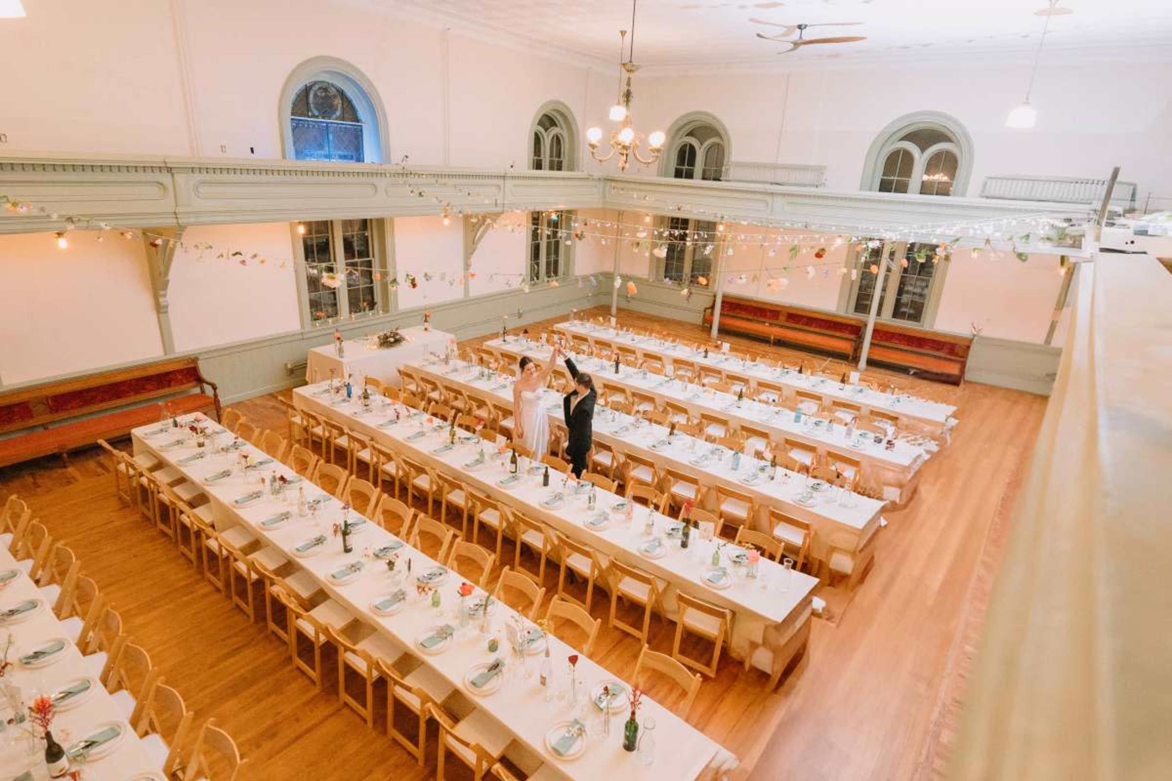 An elegantly arranged banquet hall features long tables decorated with floral centerpieces and string lights overhead, while a couple stands together at one end.