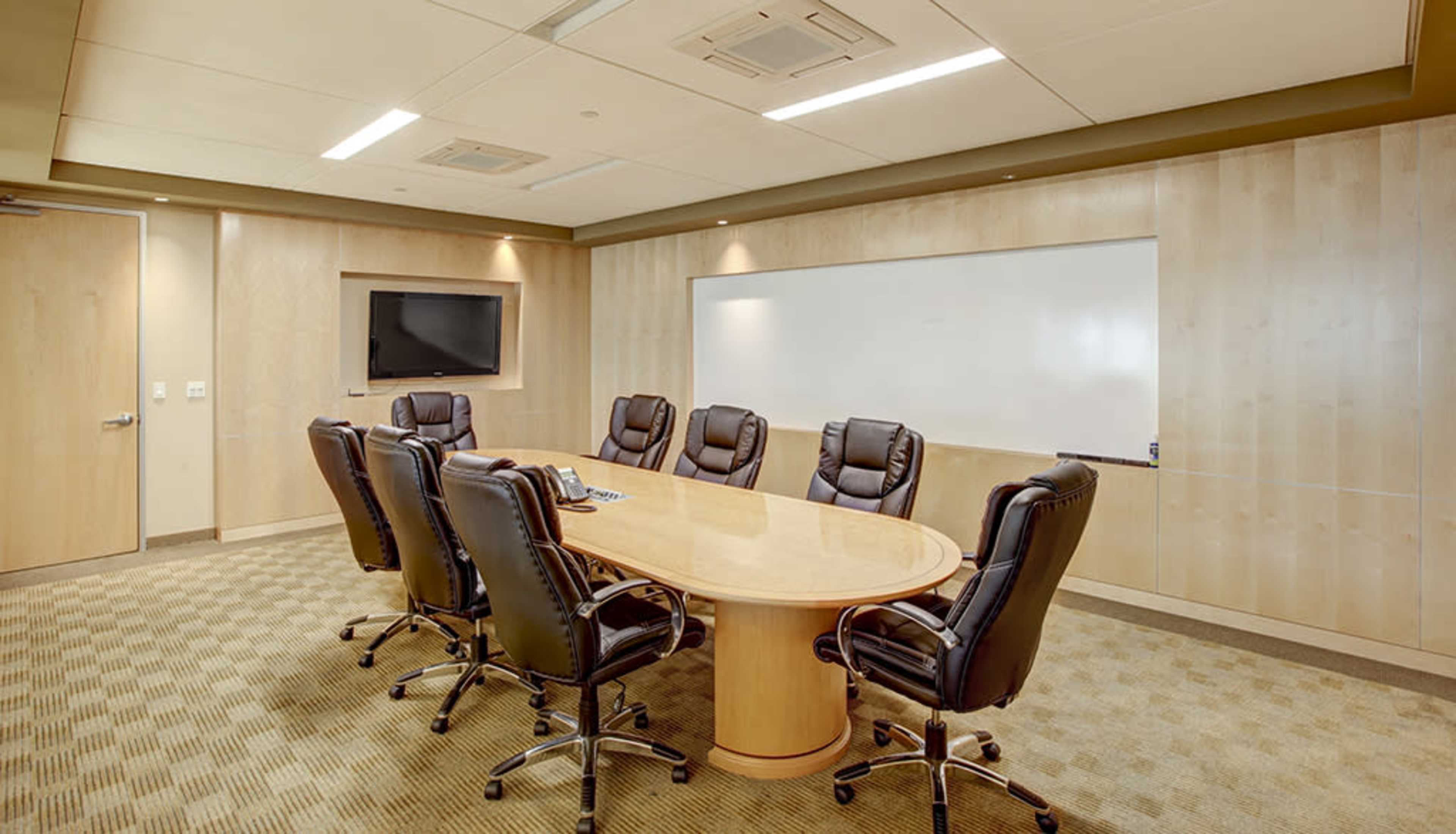 A modern conference room features a large oval table surrounded by eight executive chairs, with a whiteboard and a TV mounted on the wall.