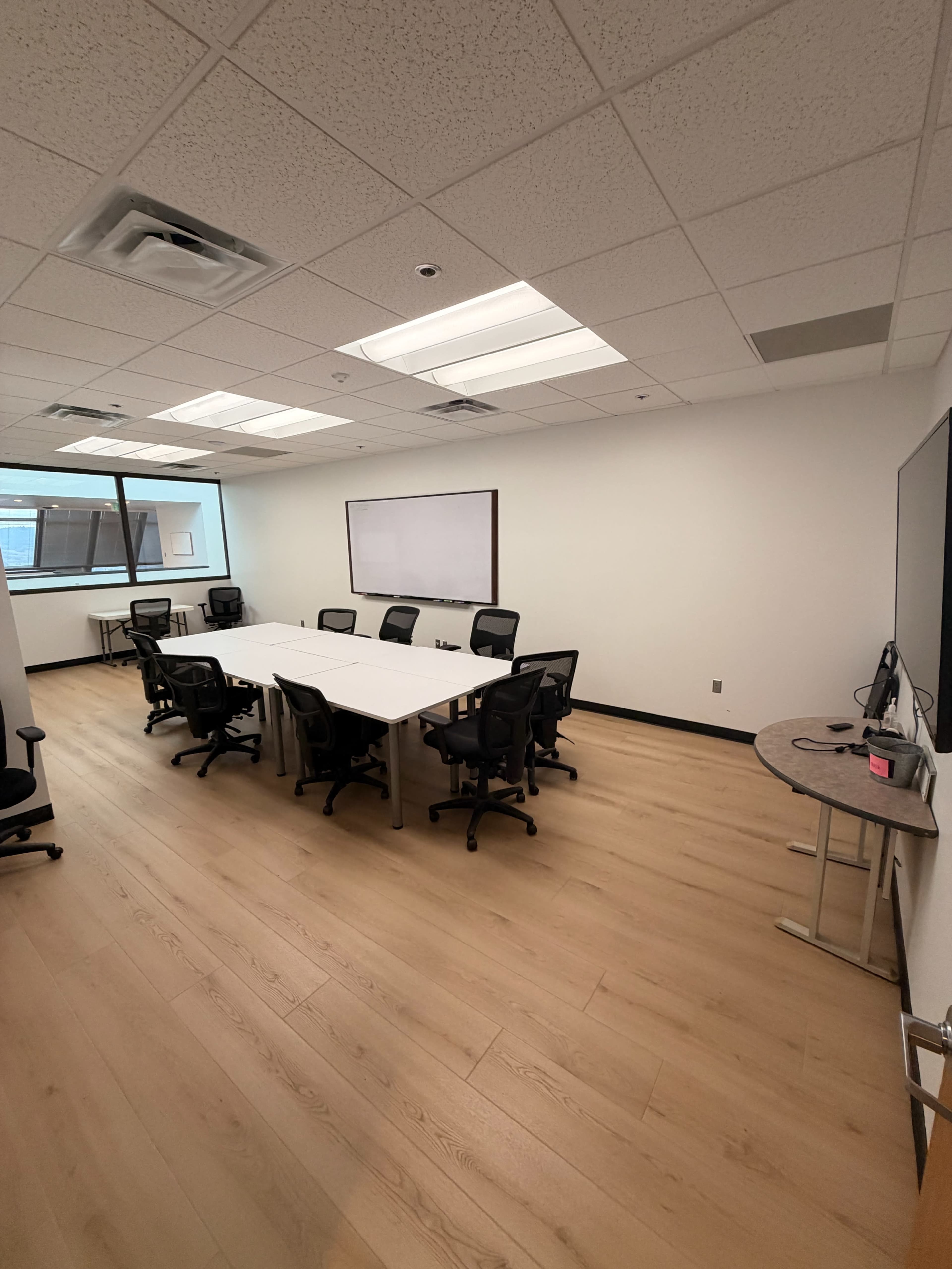 The image shows a modern conference room featuring a large table surrounded by black office chairs, with windows allowing natural light and a whiteboard on one wall.