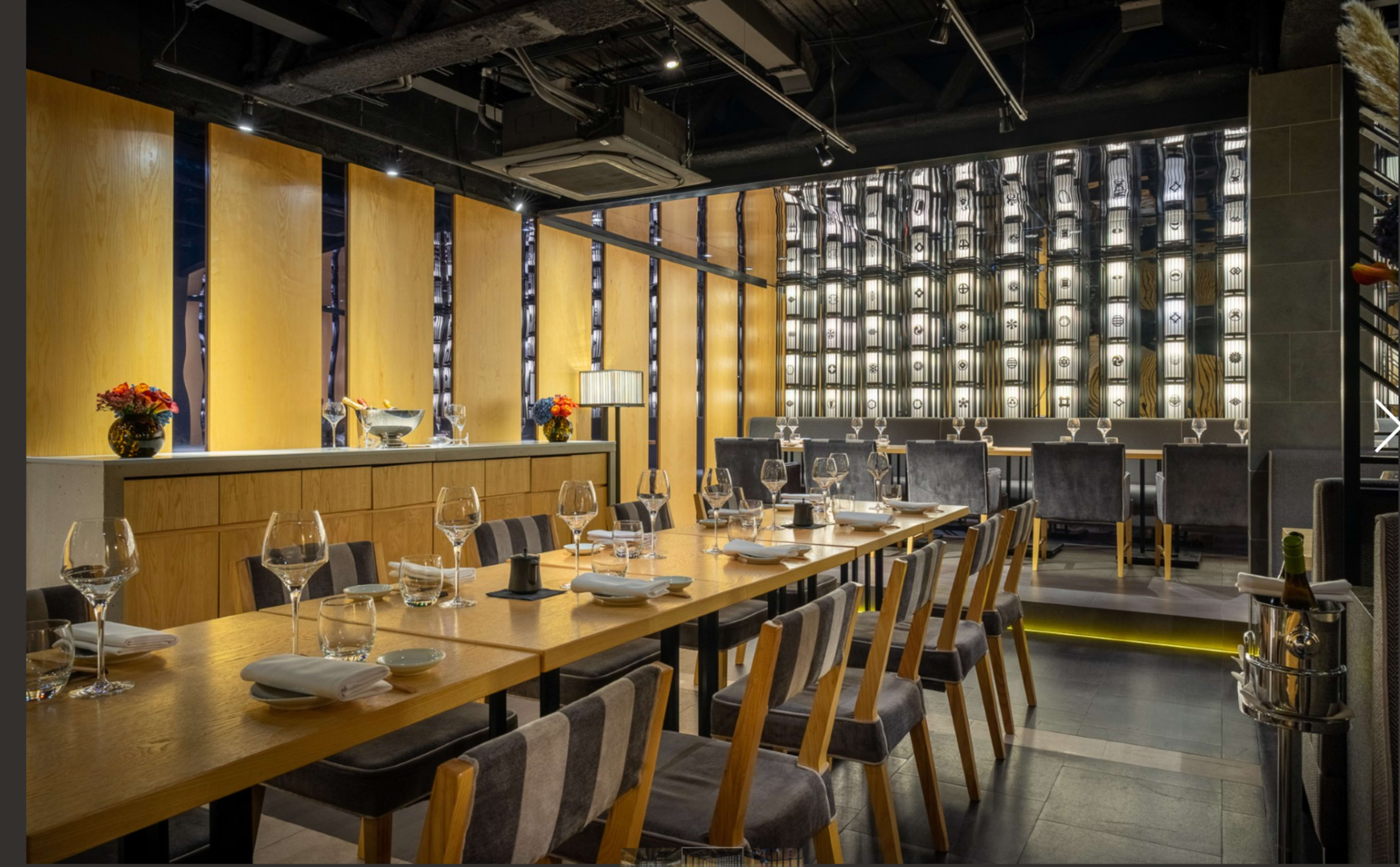 The image shows an upscale restaurant interior featuring a long wooden table set with glassware and a wall adorned with shelves of bottles.