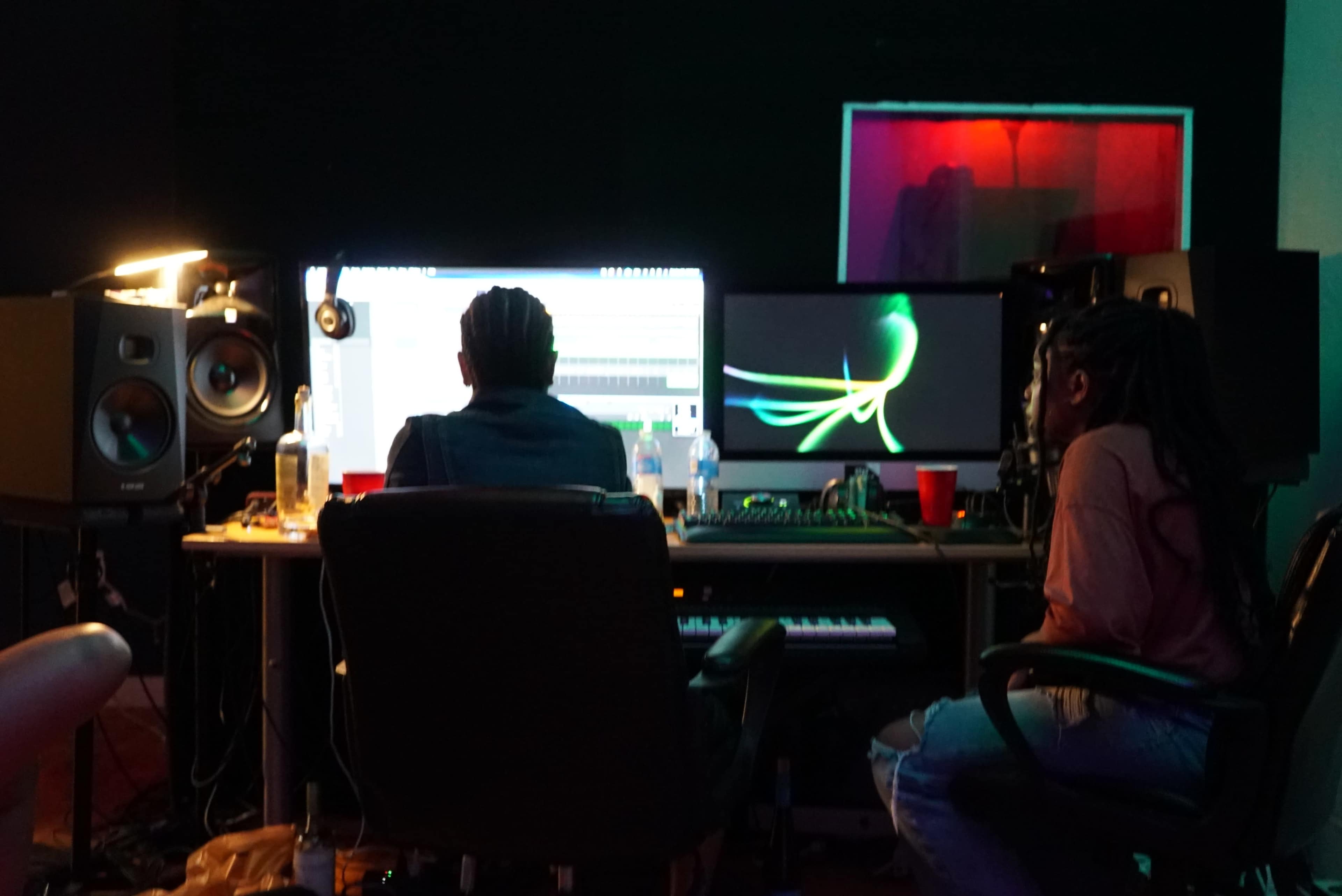 Two individuals work in a recording studio, focused on computer screens displaying audio software and visual graphics, with equipment and drinks nearby.