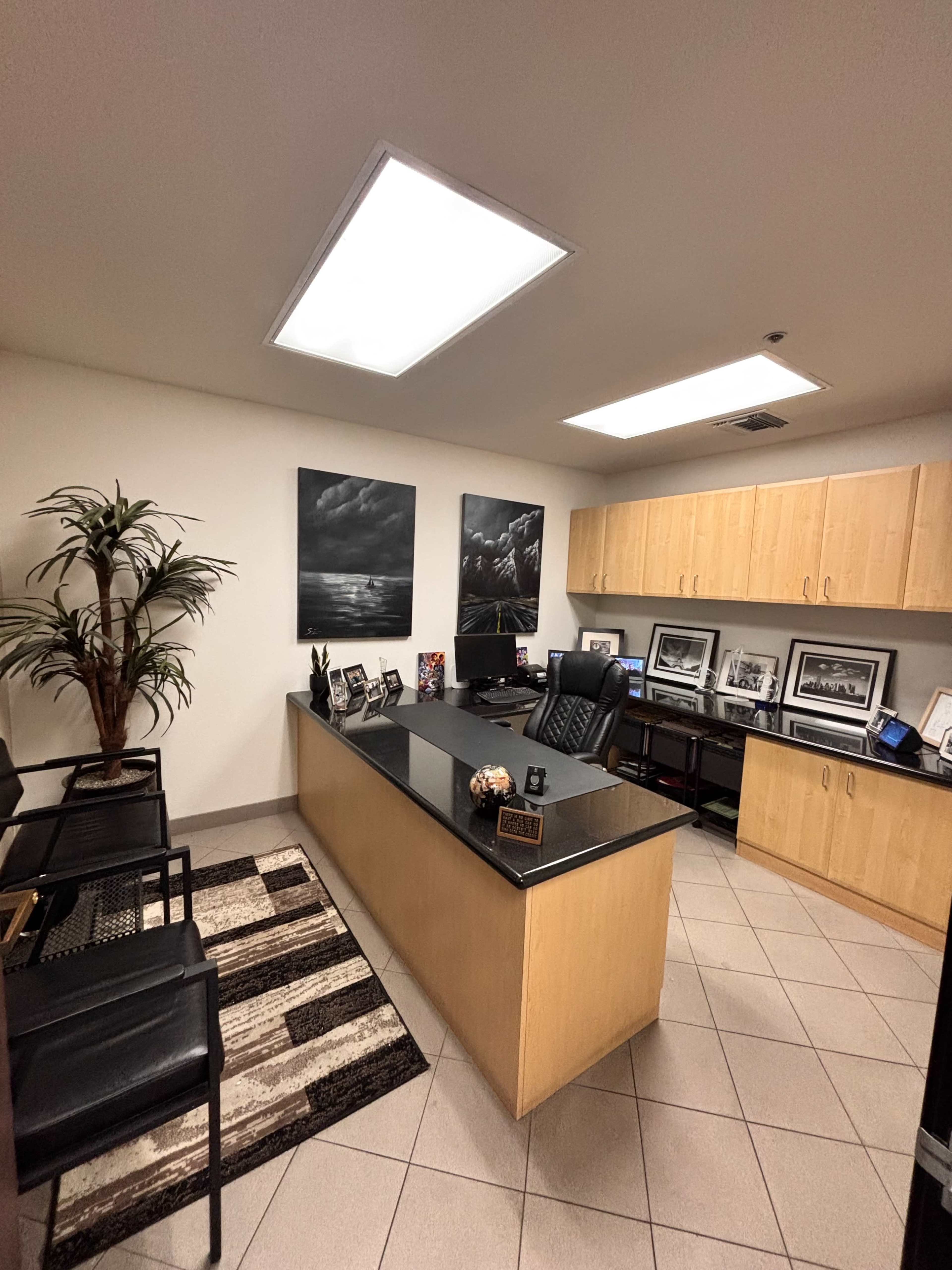 The image shows a modern office space with a black desk, wooden cabinets, photographs on the walls, and a potted plant in the corner.