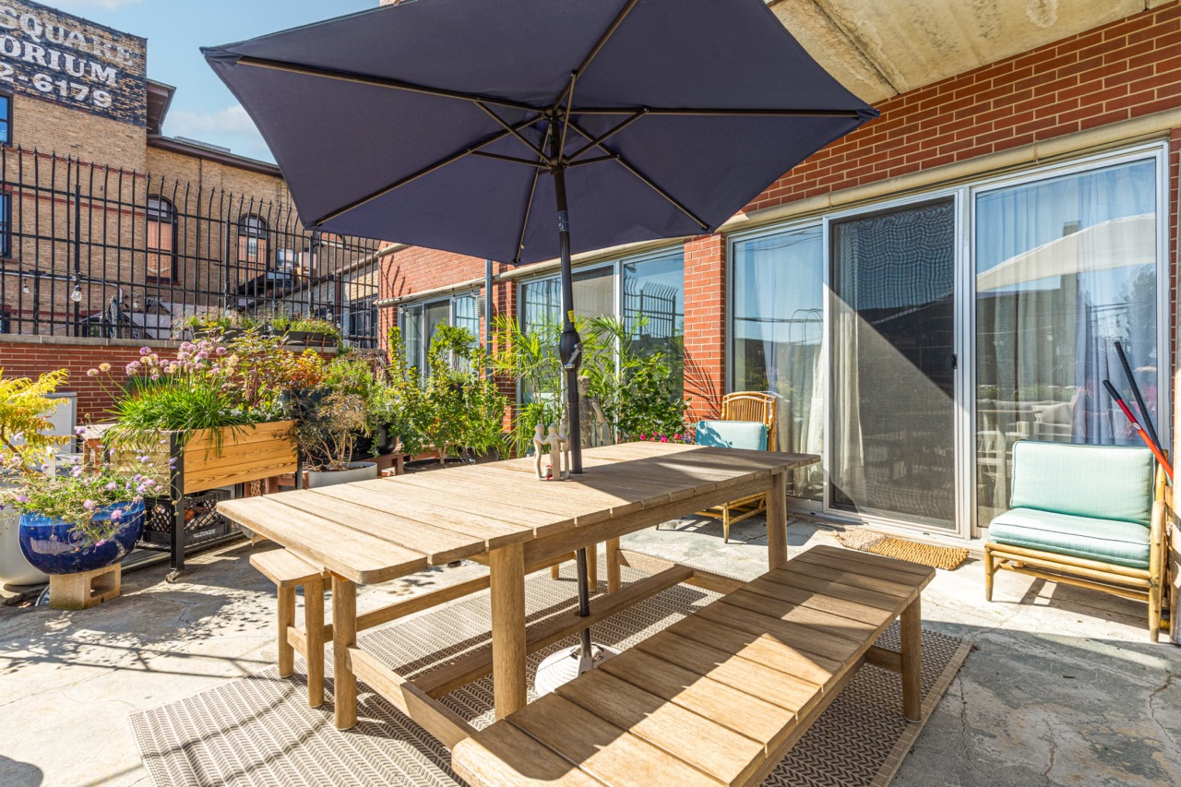 A wooden dining table with benches is shaded by a large umbrella on a terrace surrounded by potted plants and buildings.