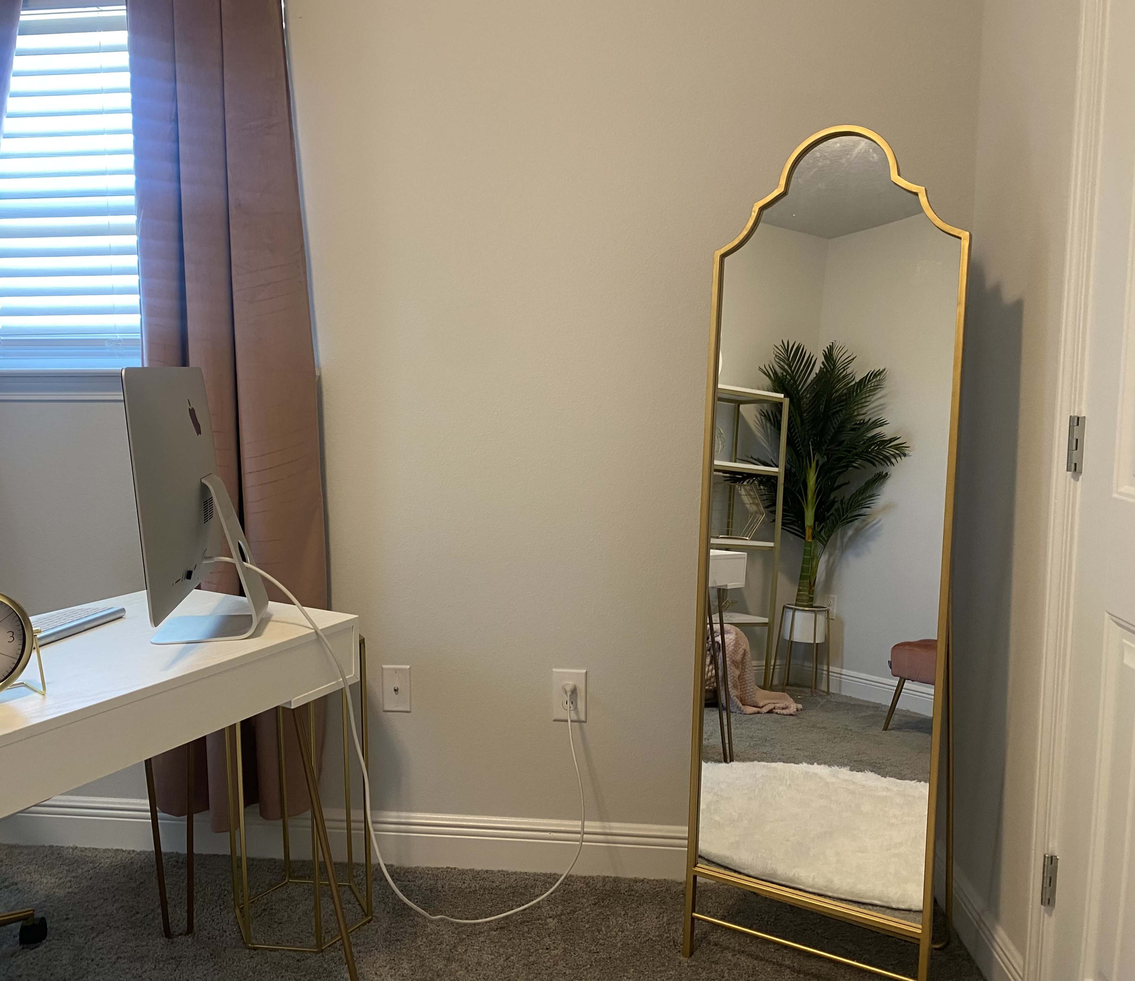 A styled corner of a room featuring a tall, gold-framed mirror beside a white desk with a computer, a potted plant, and a soft rug on the floor.