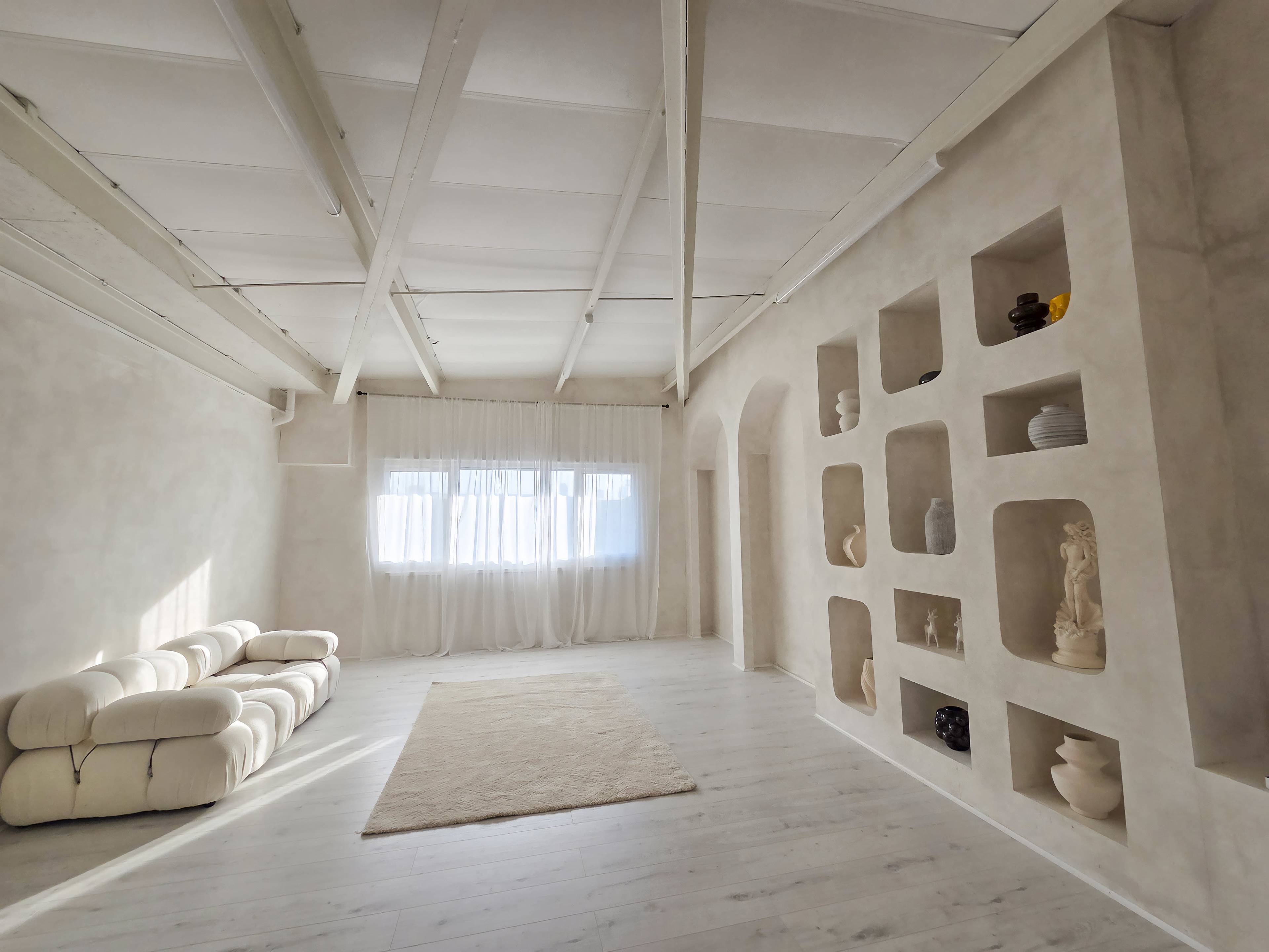 The image shows a bright, minimalist living room with a white sofa, light-colored wooden flooring, and decorative niches in the walls displaying various sculptures and vases.