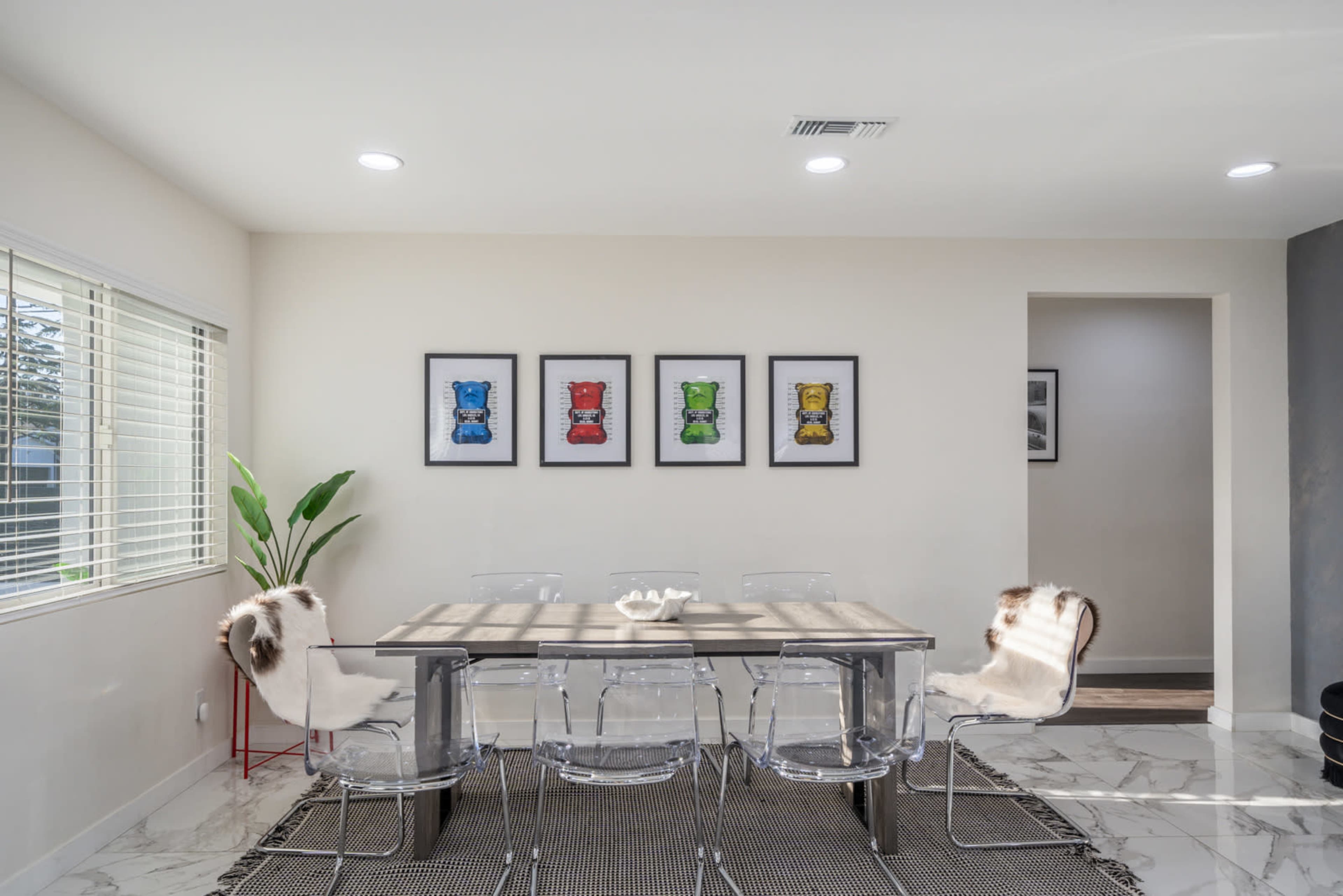 A modern dining area features a rectangular table surrounded by transparent chairs, complemented by framed artwork on the wall and a window allowing natural light.