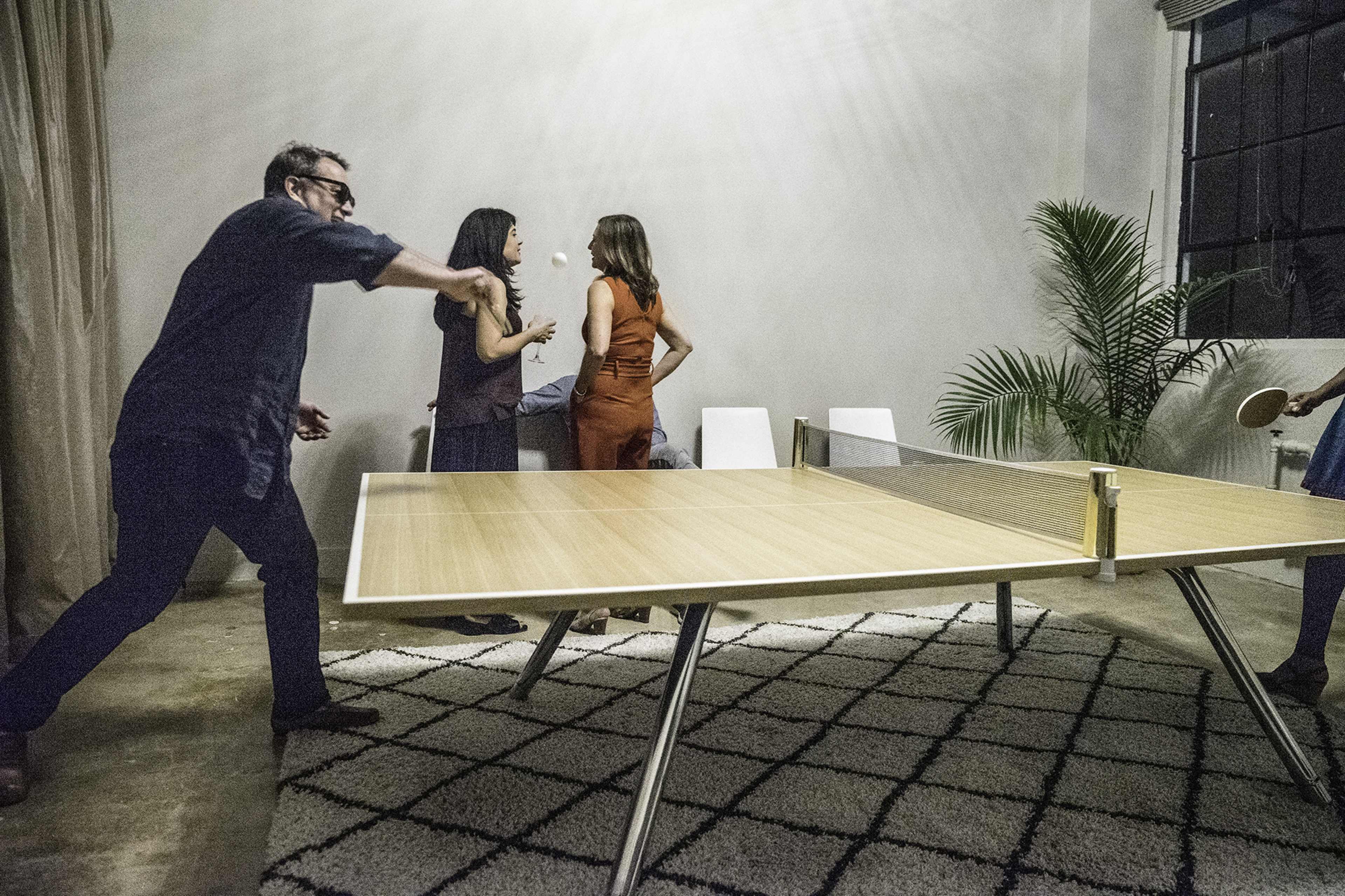 Four people are playing ping pong around a large table in a well-lit room with a rug on the floor.
