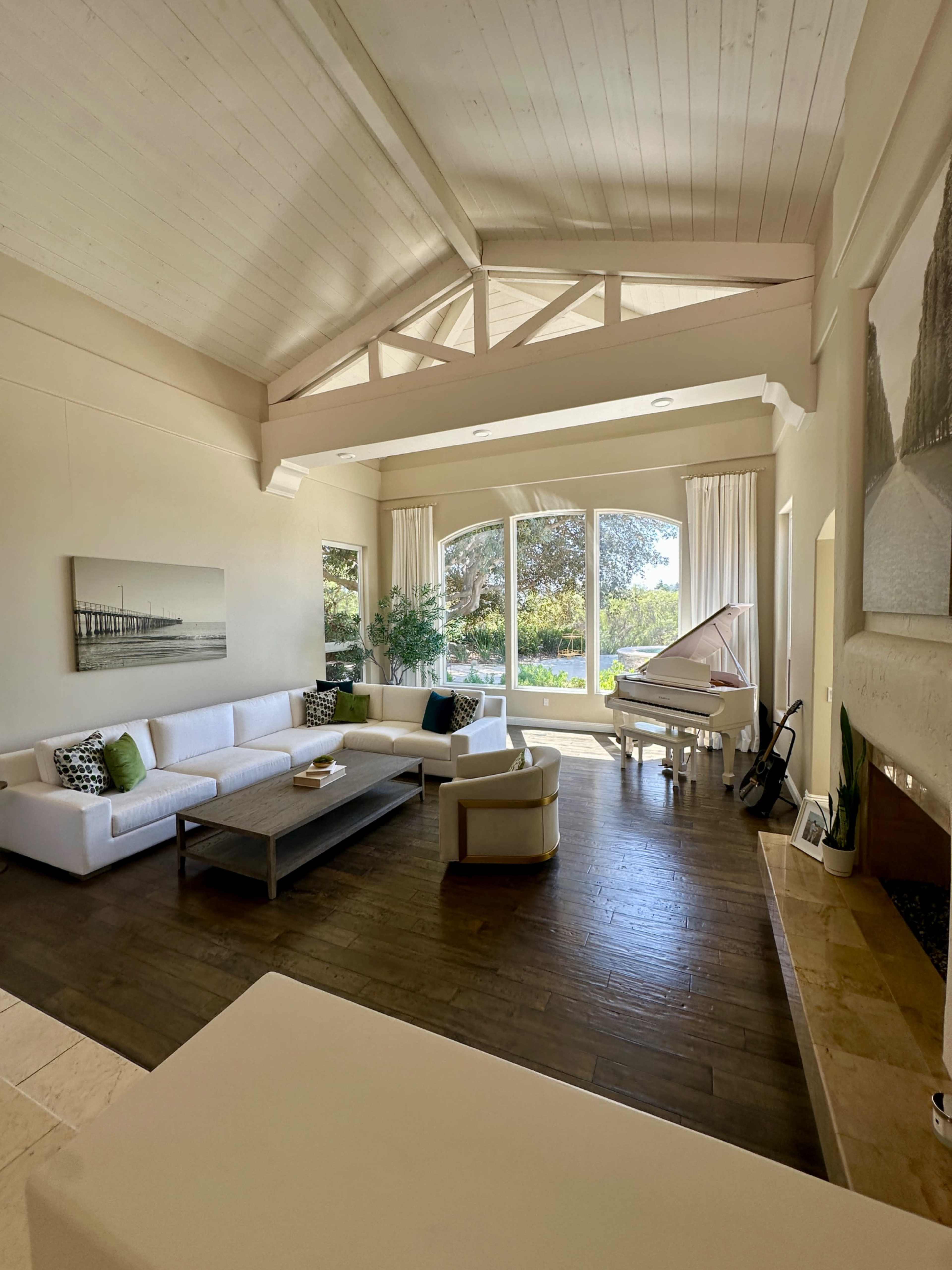 The spacious living room features large windows with natural light, a white sectional sofa, a grand piano, and a view of the outdoors.