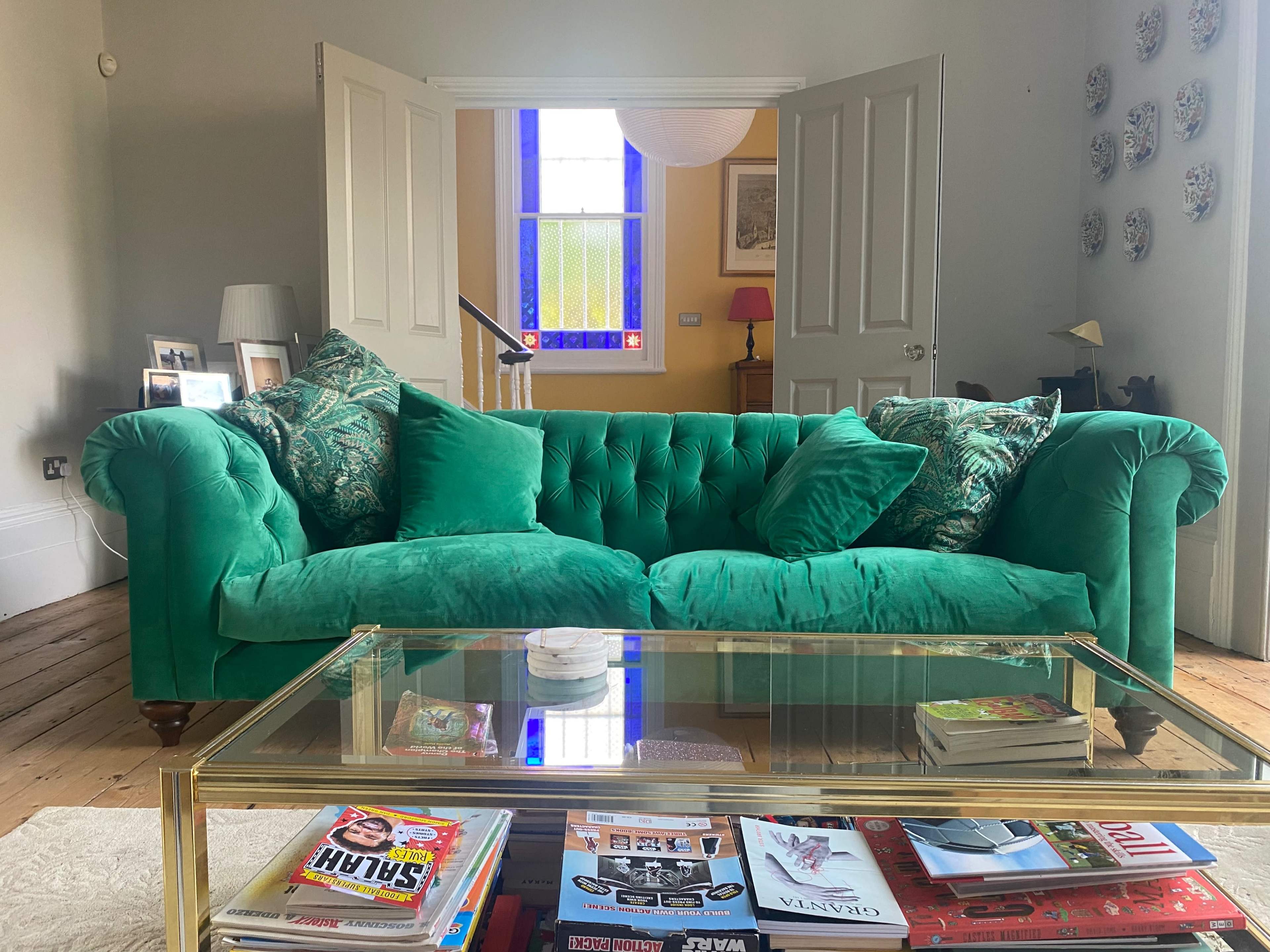 A green tufted sofa in a living room with a glass coffee table and a bright window visible in the background.