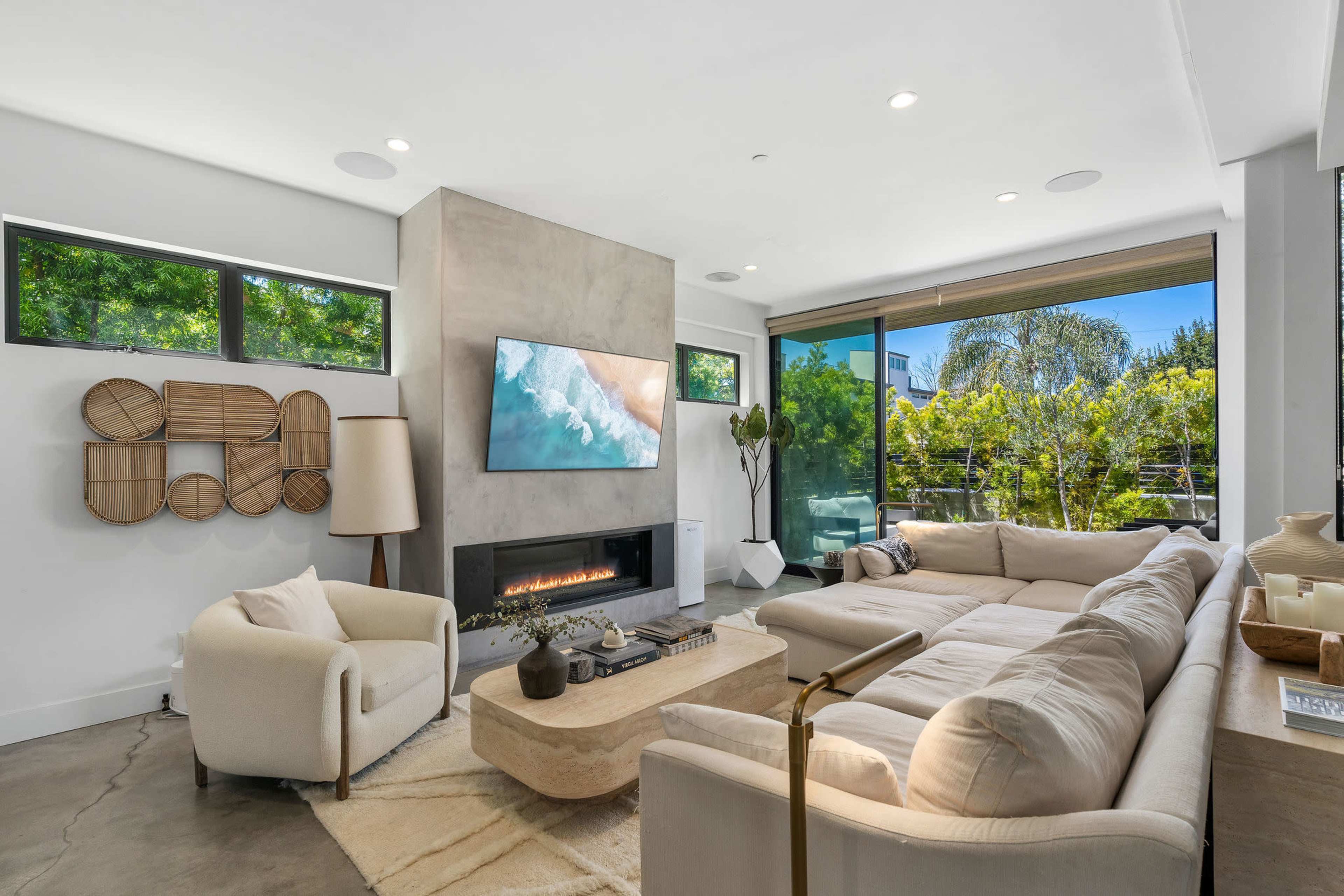 The living room features a modern design with a large sectional sofa, a minimalist coffee table, and a wall-mounted television above a sleek fireplace, all surrounded by large windows letting in natural light.