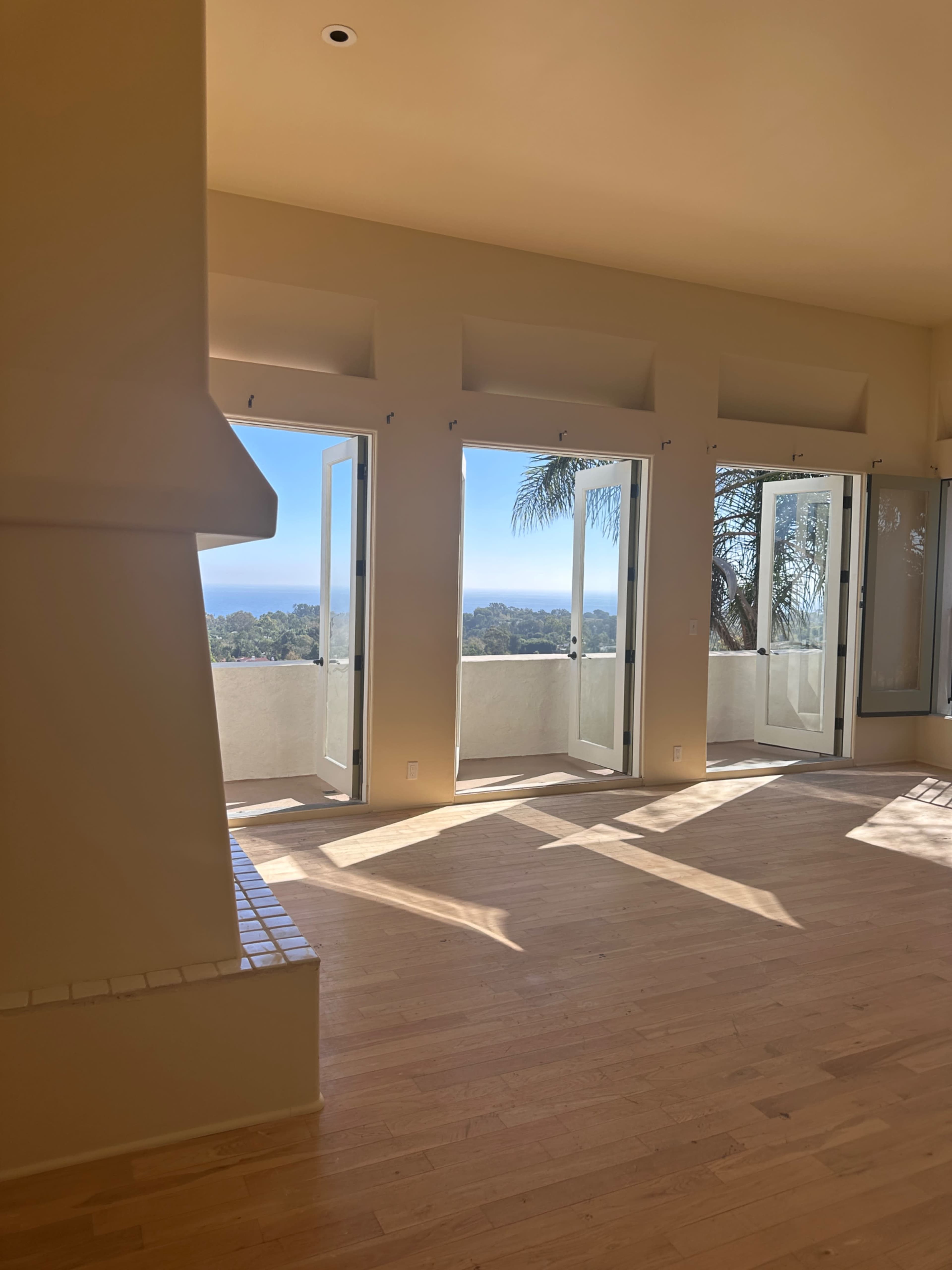 The image shows a spacious, sunlit room with multiple open French doors leading to a balcony that overlooks a scenic view.