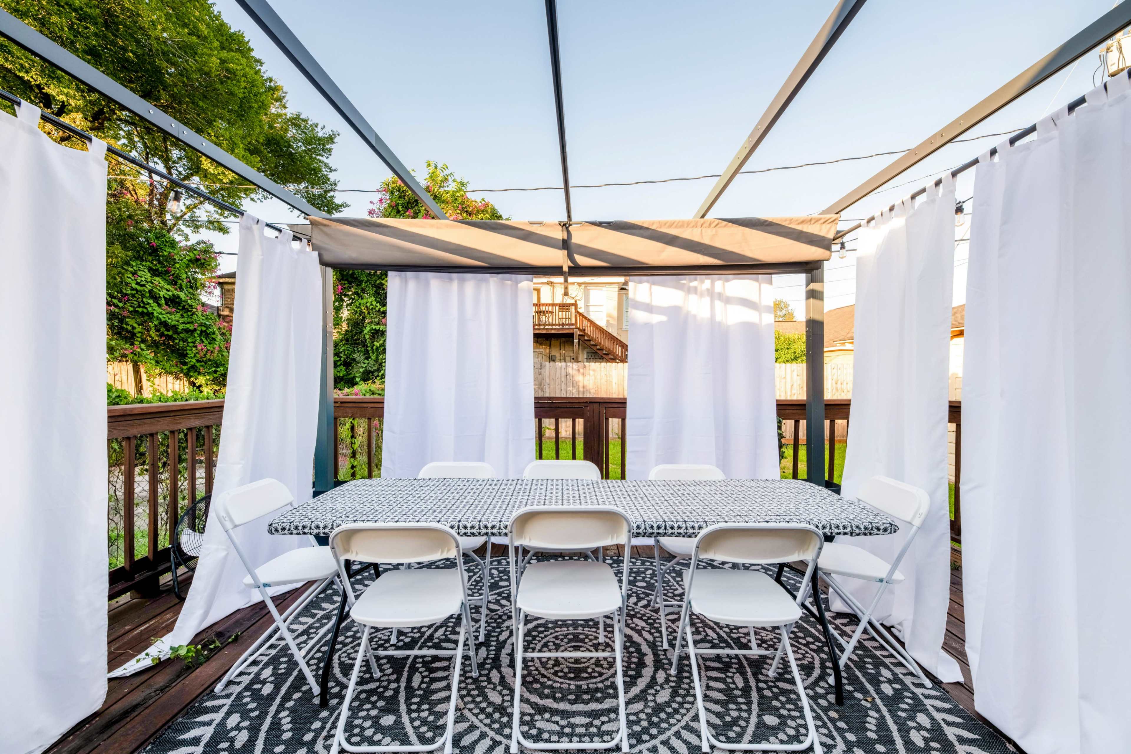 A large outdoor dining area is set with a patterned tablecloth and surrounded by white curtains and folding chairs on a wooden deck.