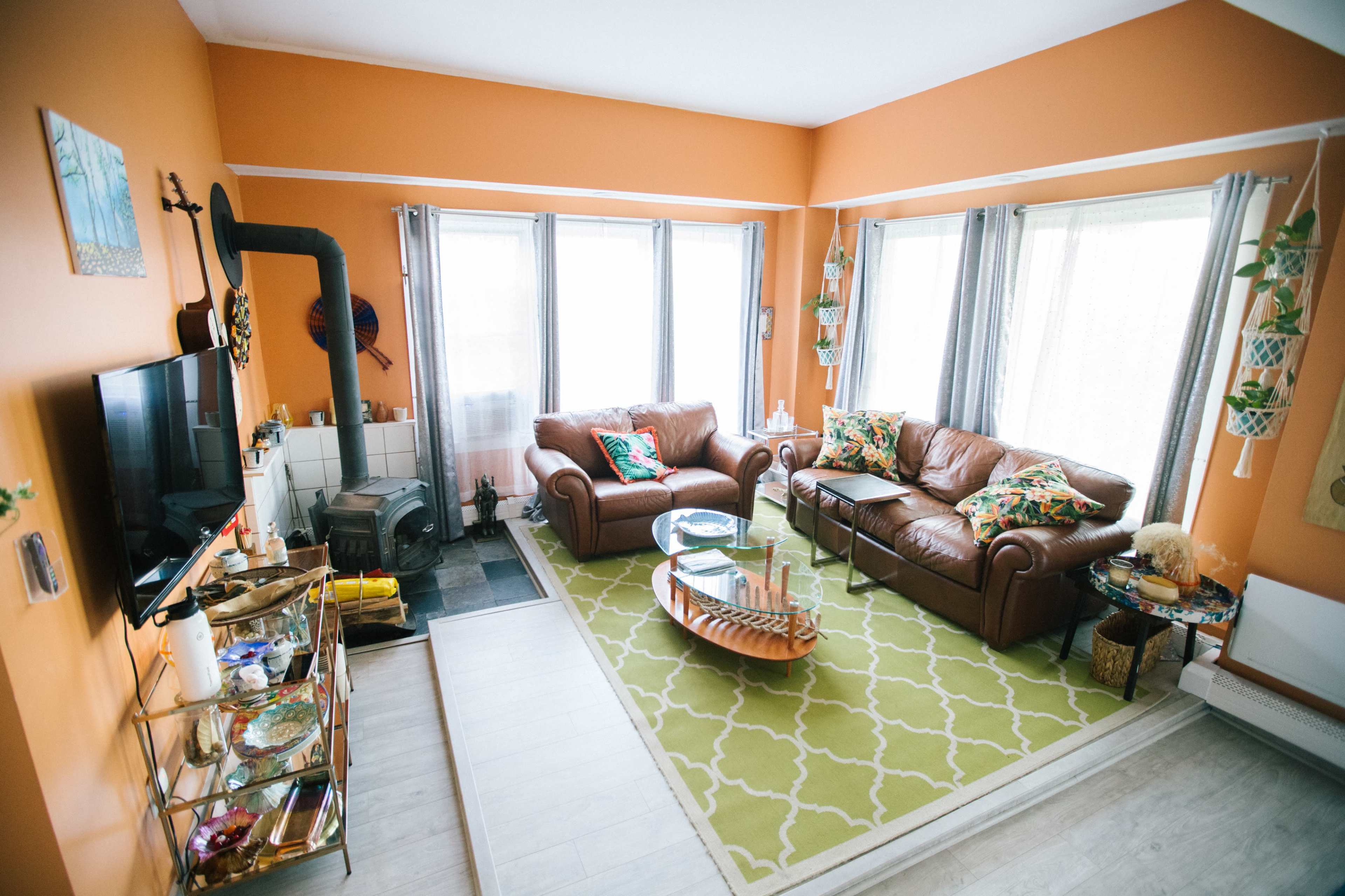 The image shows a bright living room with orange walls, two brown leather sofas, large windows, and a patterned area rug.
